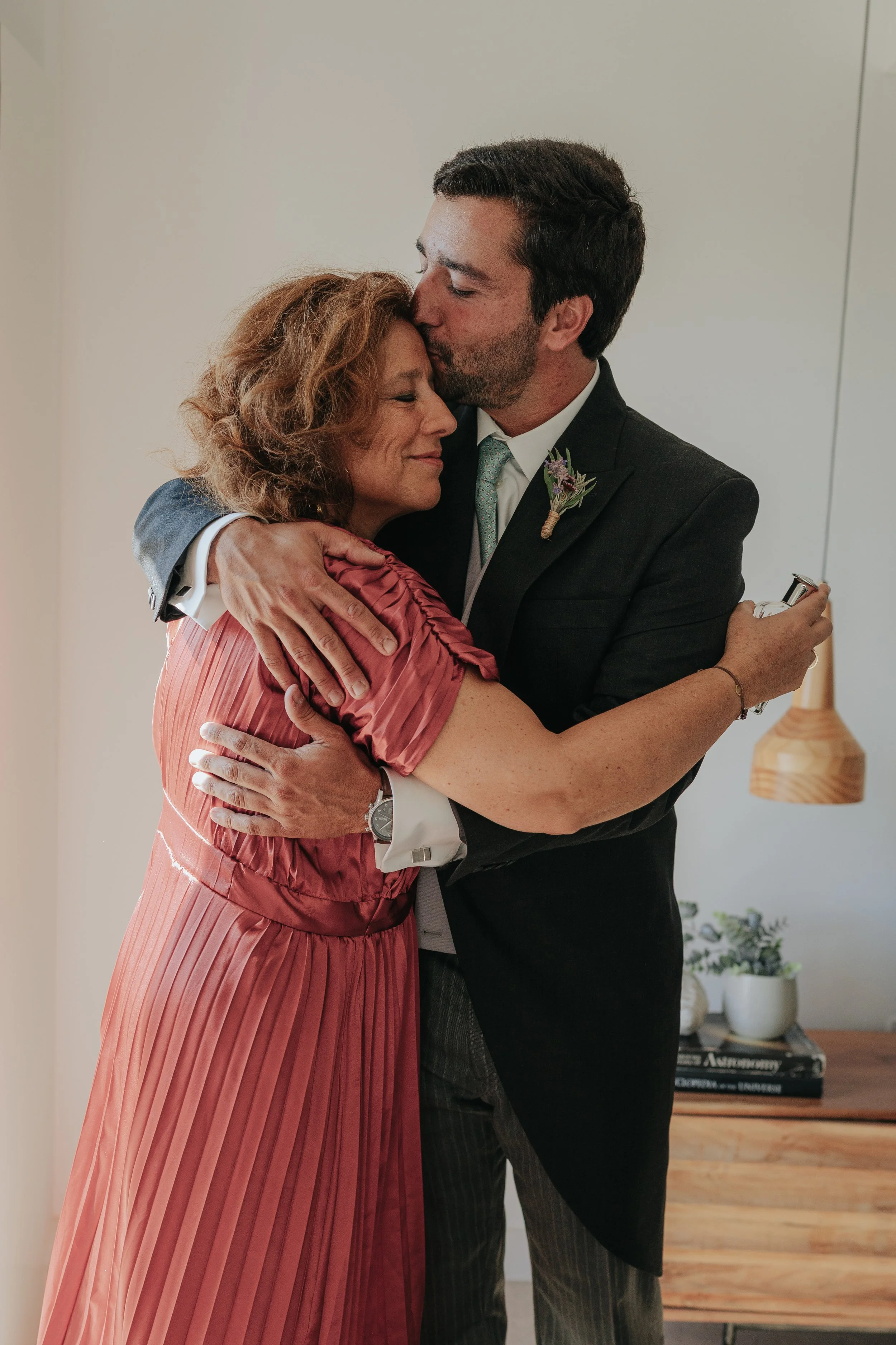 A man in a suit and a woman in a pink dress embrace as he kisses her forehead with a gentle smile. They appear to be sharing an intimate moment indoors.