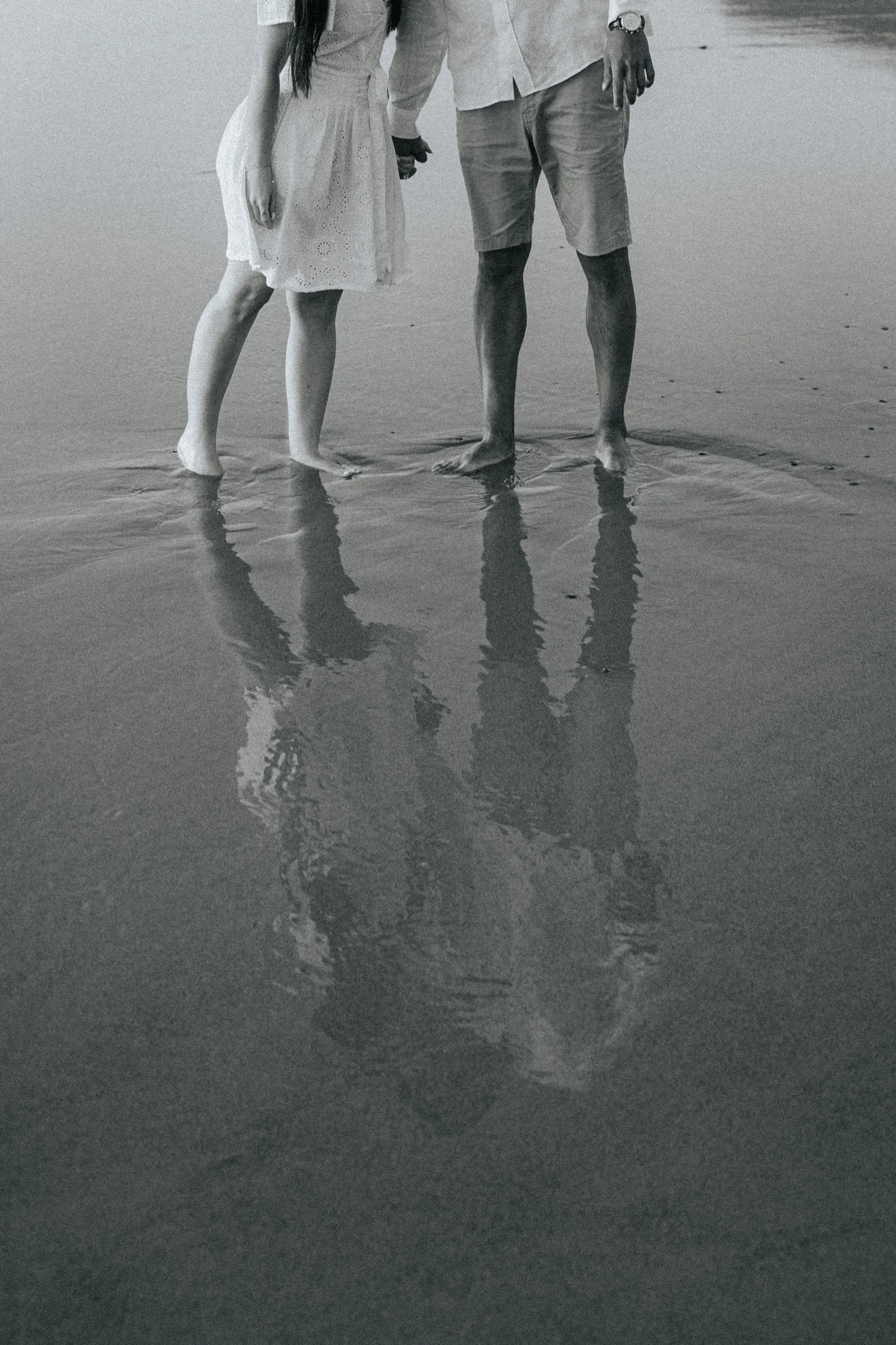 A couple holding hands on the beach with their feet in the sand, reflected in the shallow water.