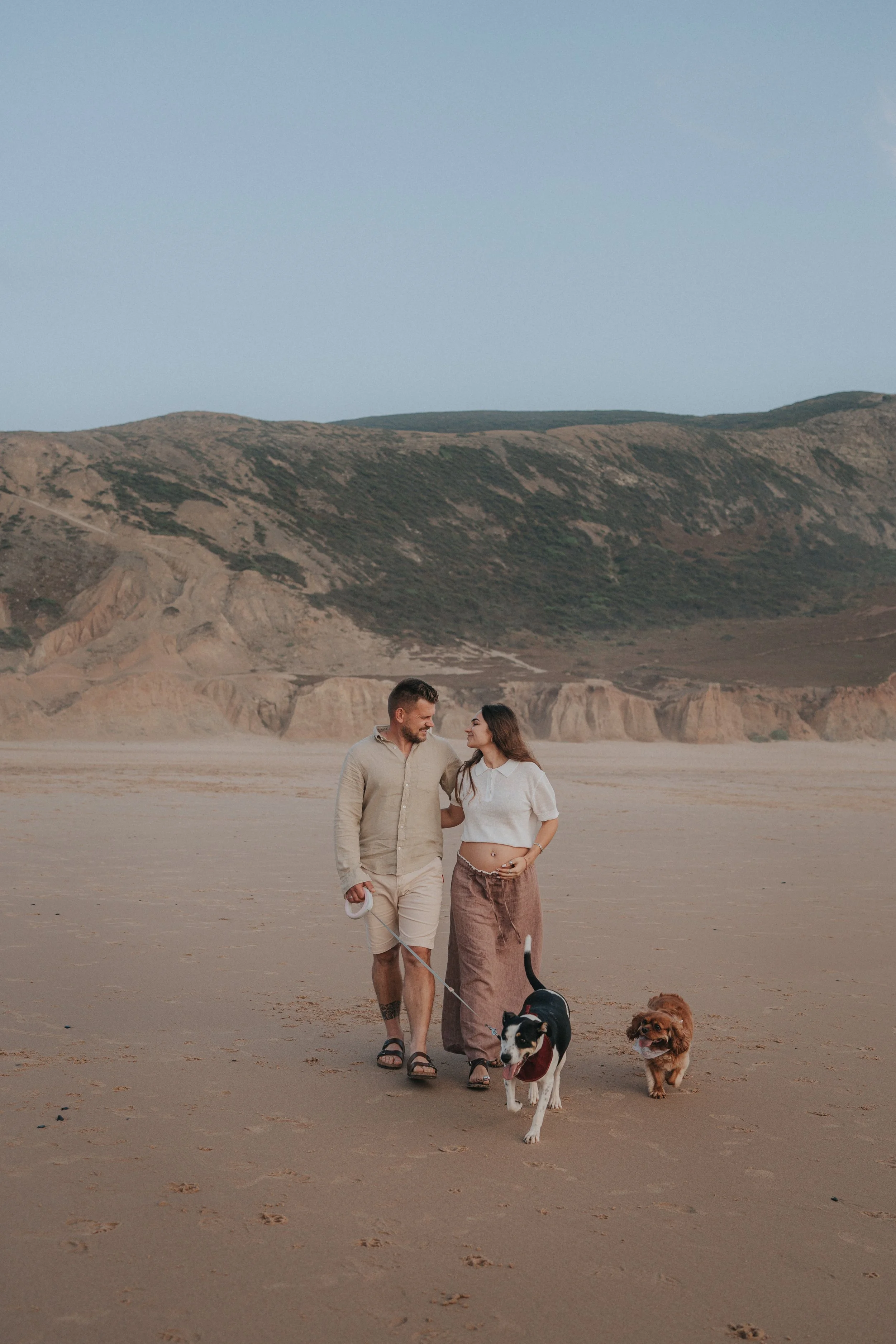 Couple walking with two dogs on a sandy beach with hills in the background.