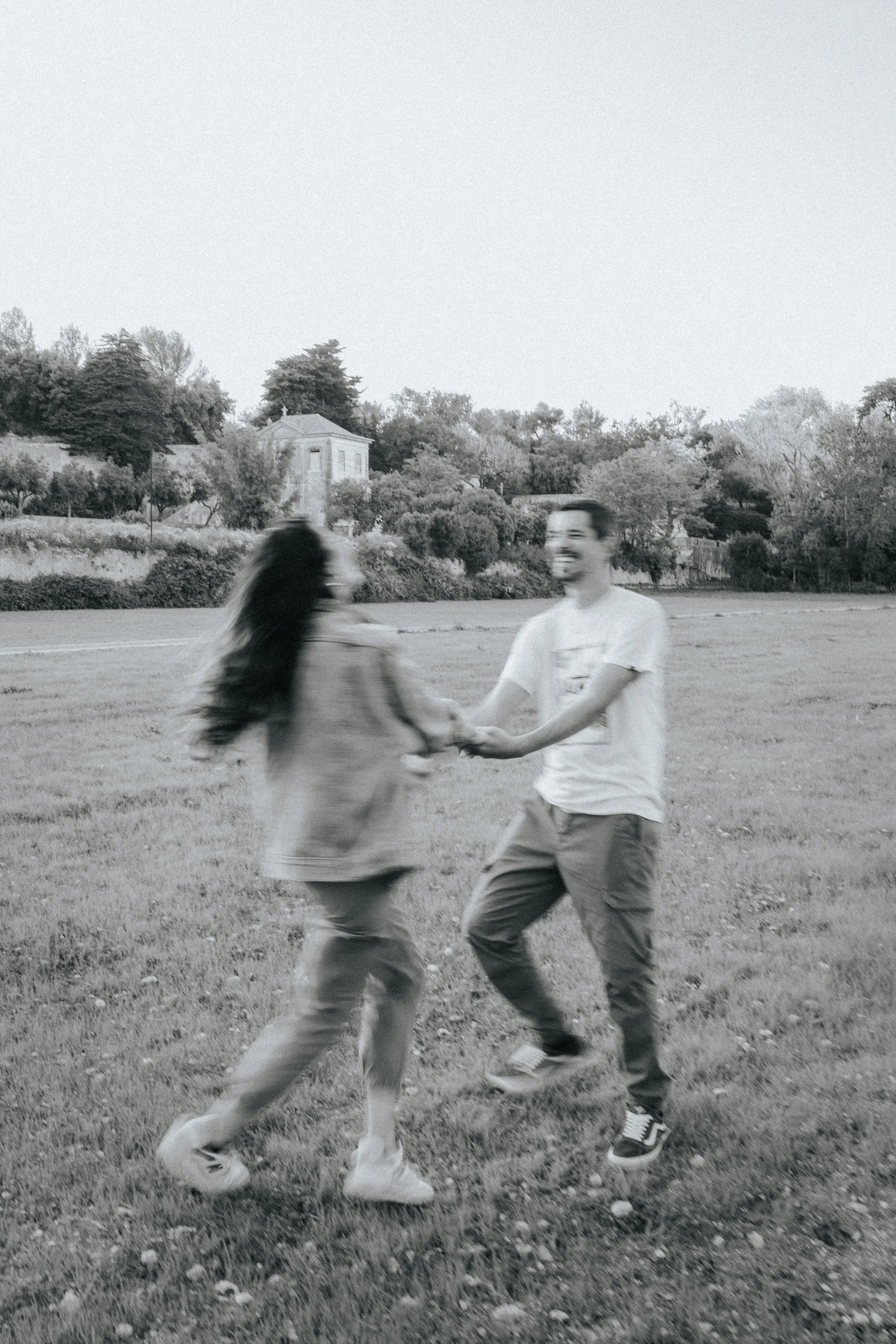 Two people are dancing or playing together in a grassy park, holding hands and smiling, with trees and a house in the background.