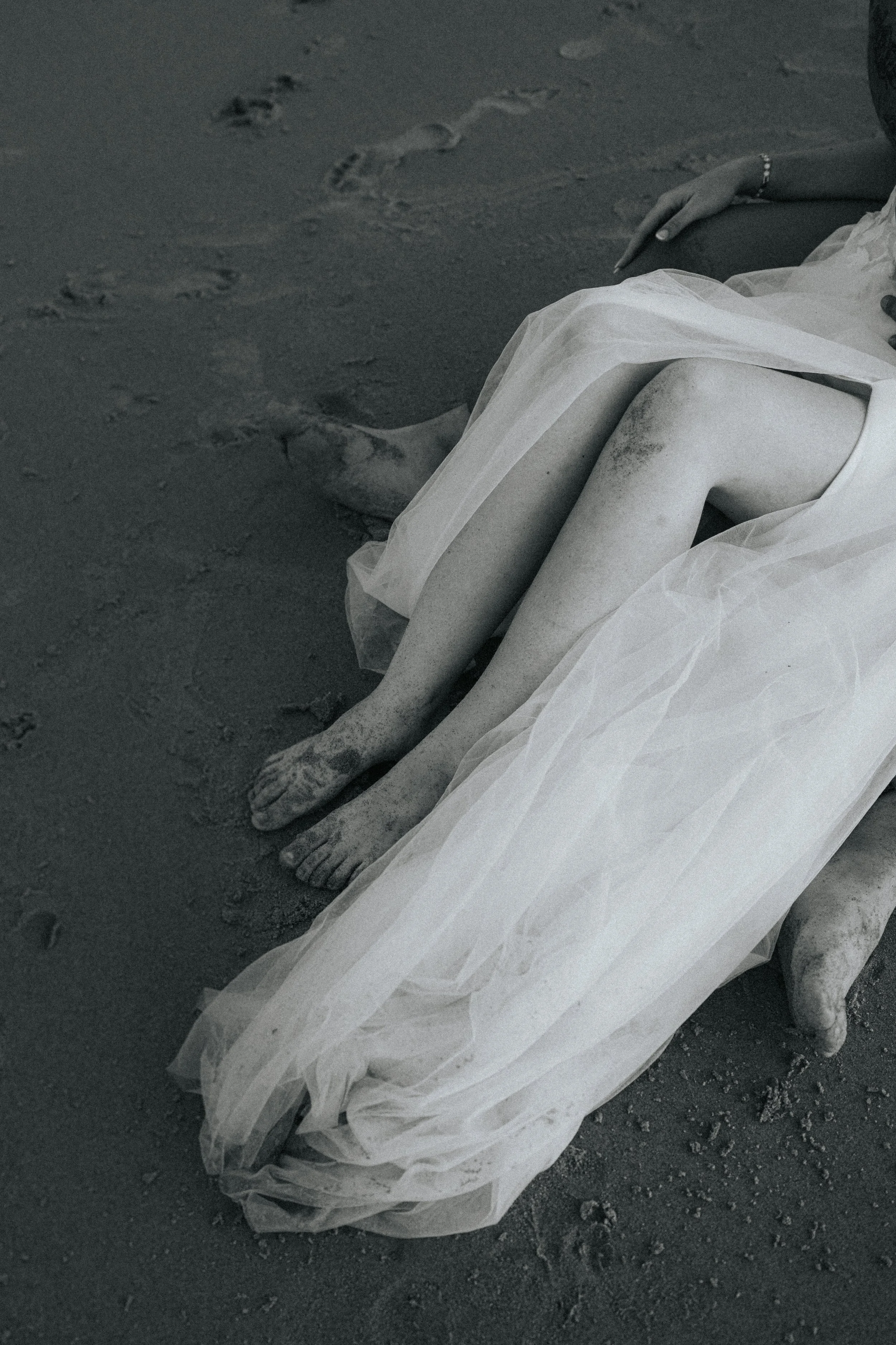 A person lying on the sand with a white veil partially covering their legs. The person's hand is resting on their knee, and there are footprints visible in the sand nearby.
