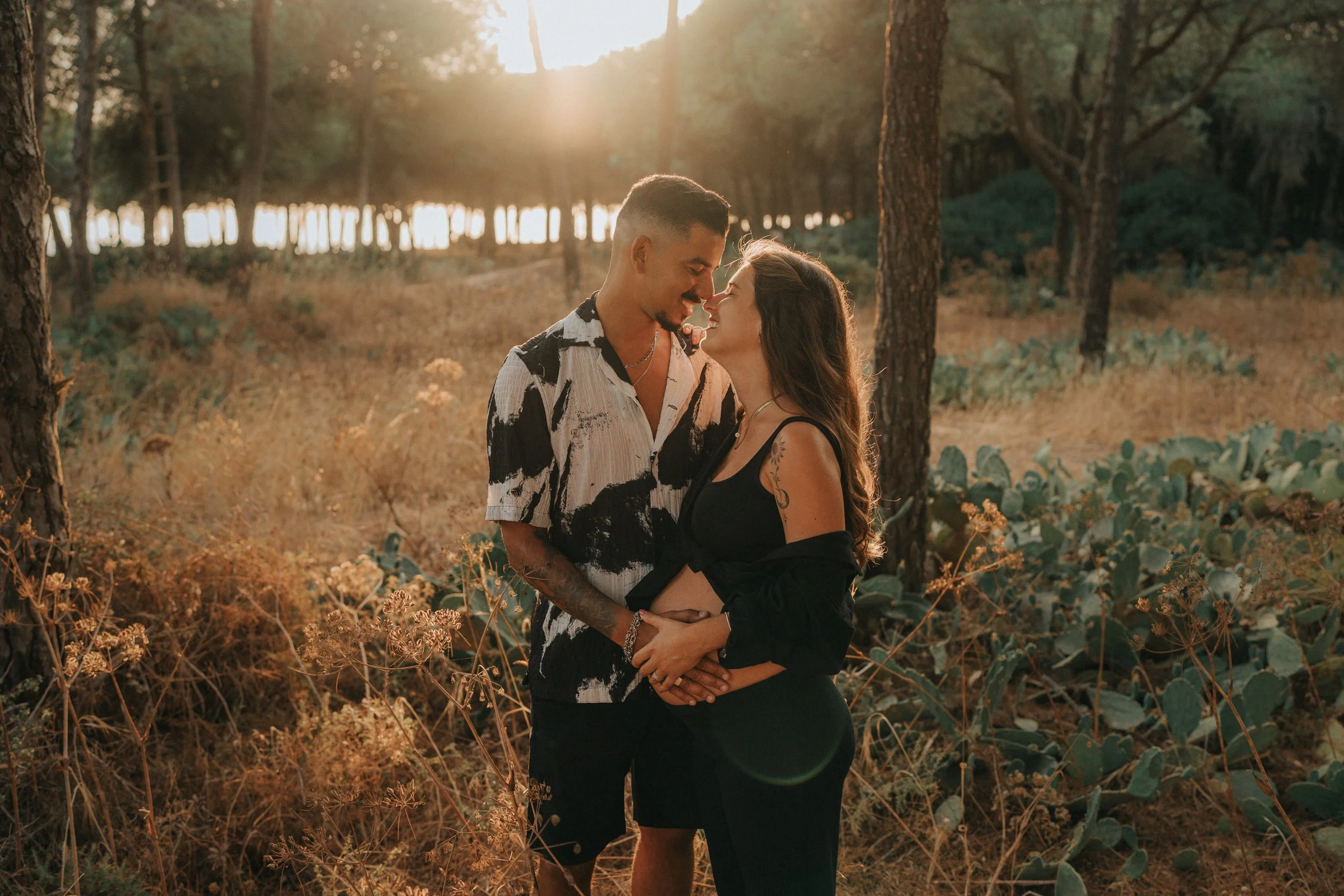 A couple standing closely together in a forest at sunset, with the sun shining through the trees in the background. They are smiling and touching foreheads, embracing affectionately.