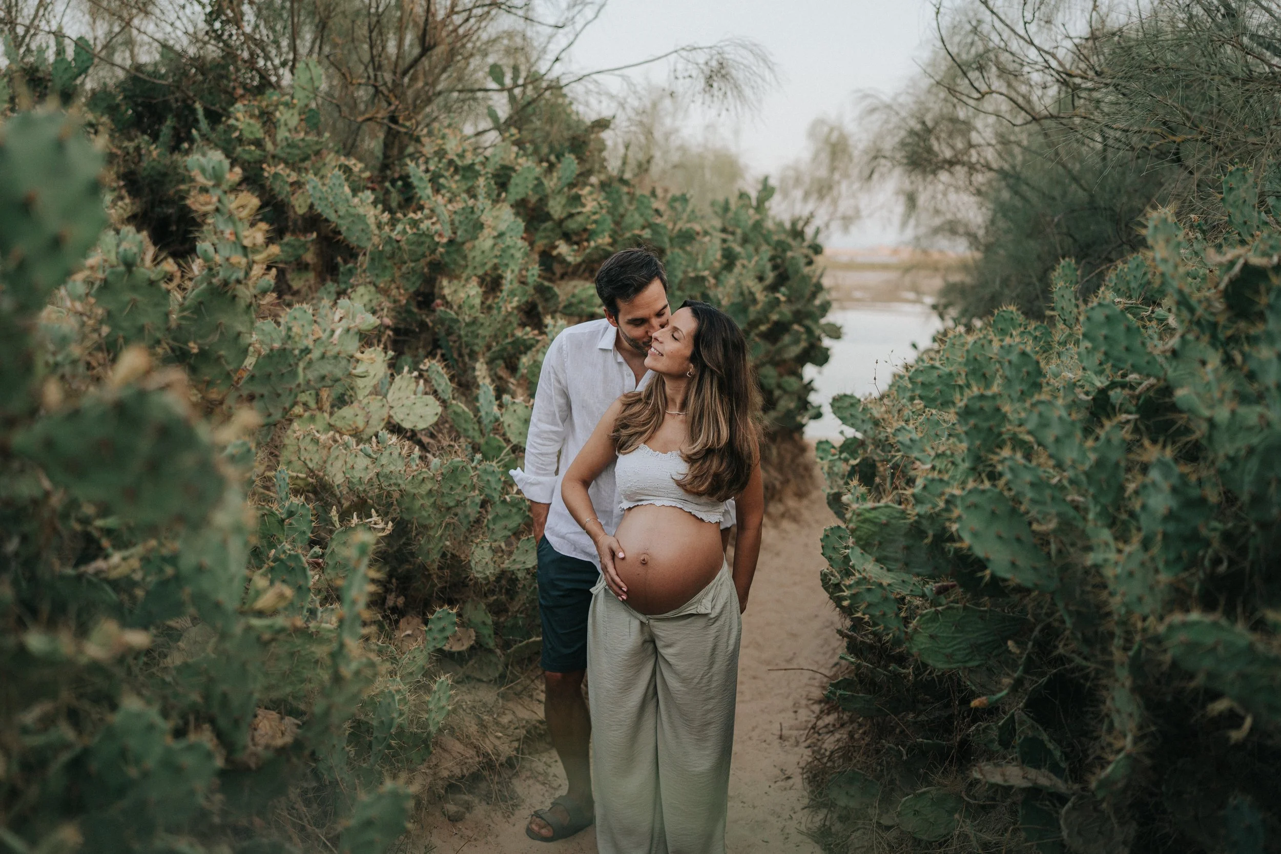 A pregnant woman and a man standing close together in a desert-like setting with cacti and dry trees, with the man kissing the woman's forehead and the woman gently touching her belly, both smiling and outdoors.