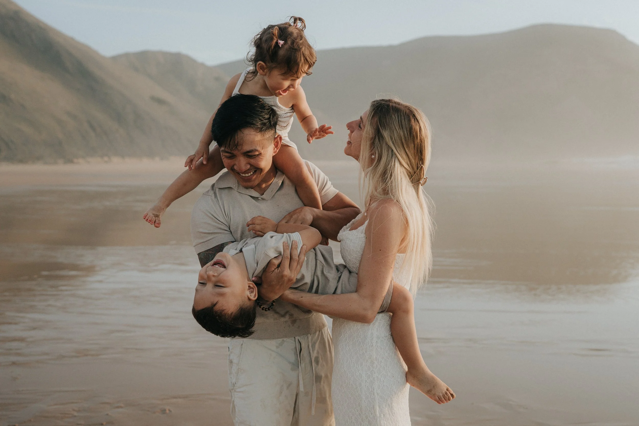 A family of four playing on the beach, with mountains in the background, during sunset. The father is holding a young boy, and the mother is standing beside them, smiling at the boy, while the girl is sitting on the father's shoulders.