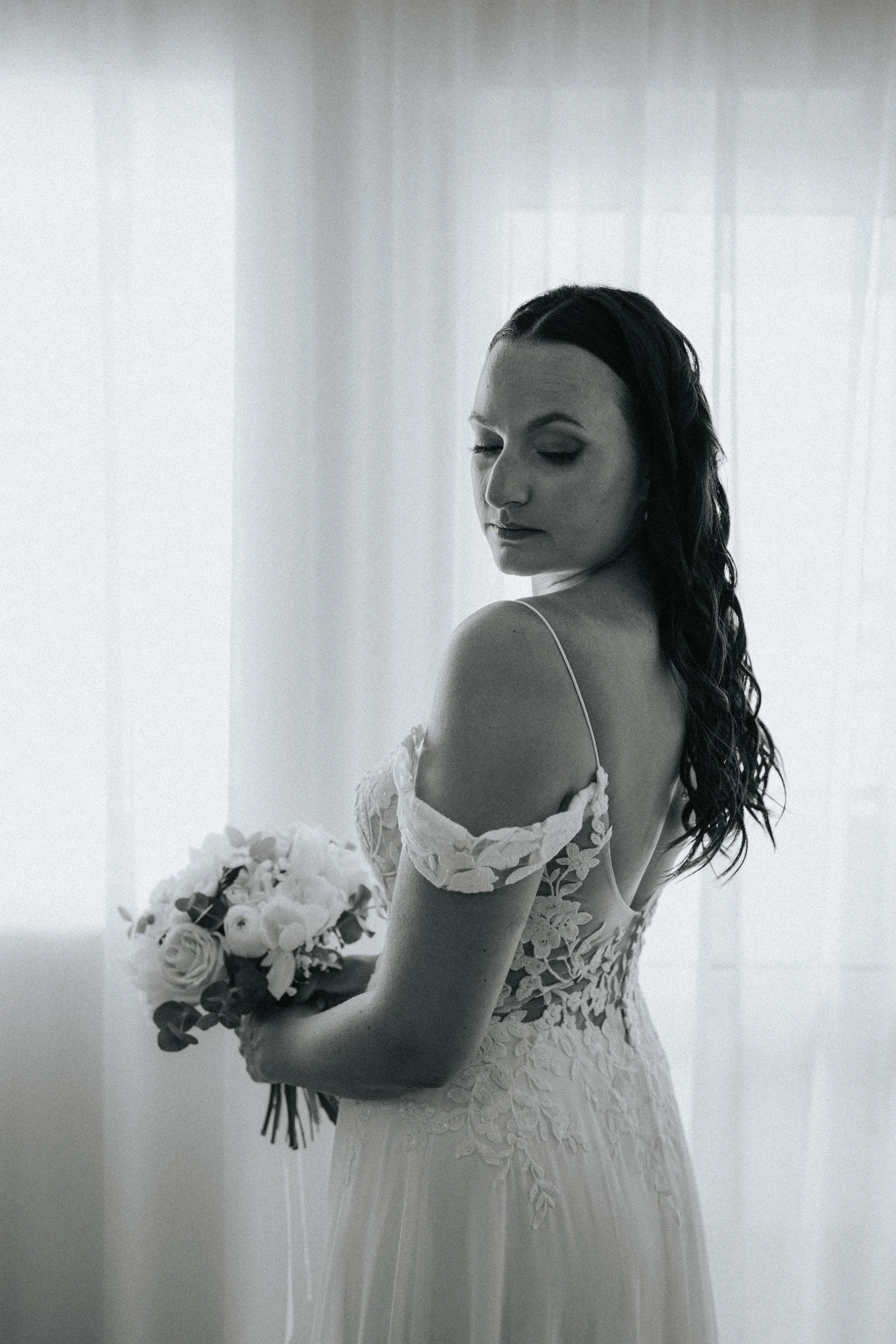A bride with long dark hair styled in waves, wearing a lace wedding dress with off-the-shoulder sleeves, holding a bouquet of white roses and greenery, standing in front of a sheer curtain with soft lighting.