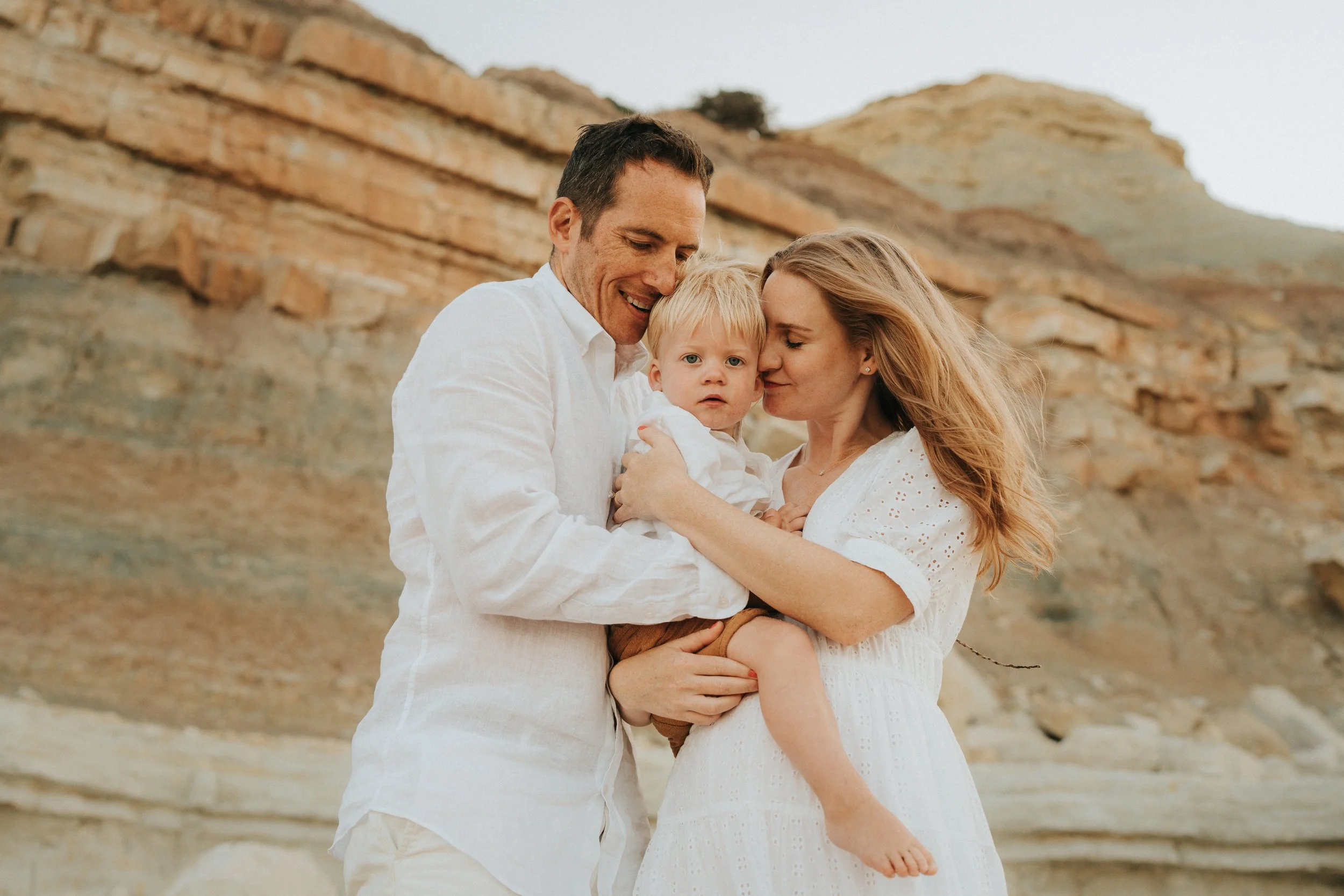 A family of three, with a man, woman, and young child, embracing each other outdoors near rocky terrain.