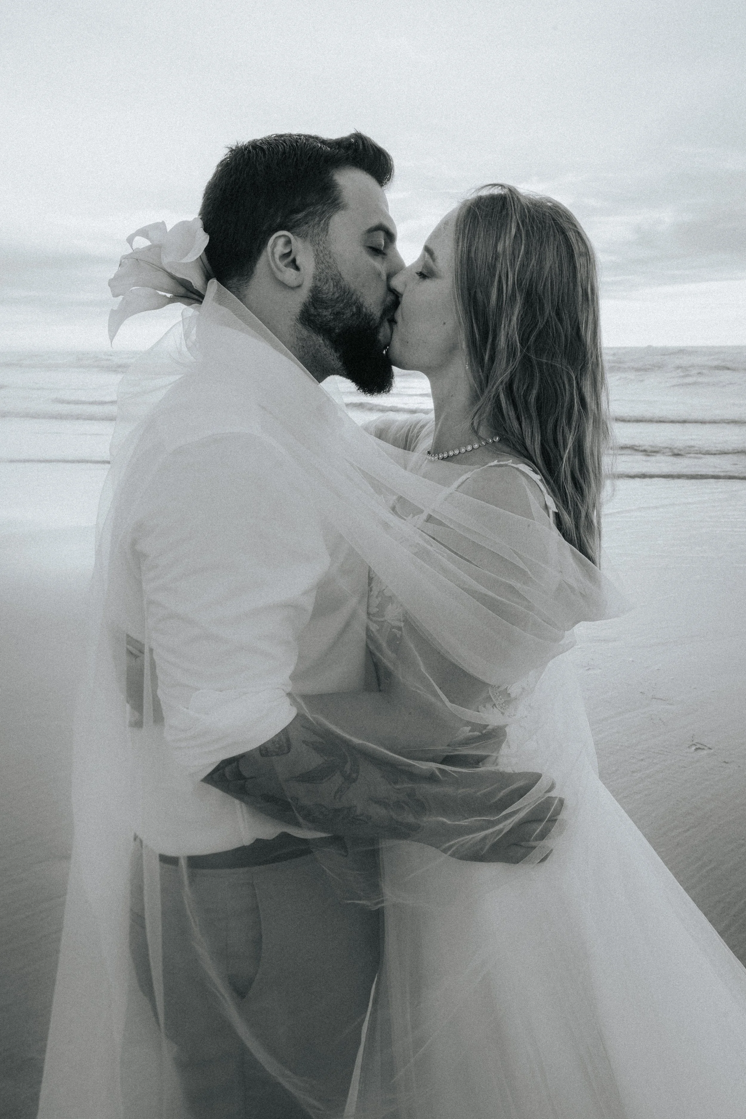 A black-and-white photo of a man and woman at the beach kissing, with the woman in a wedding dress and pearl necklace, the man in a white shirt and casual shorts, ocean waves in the background.