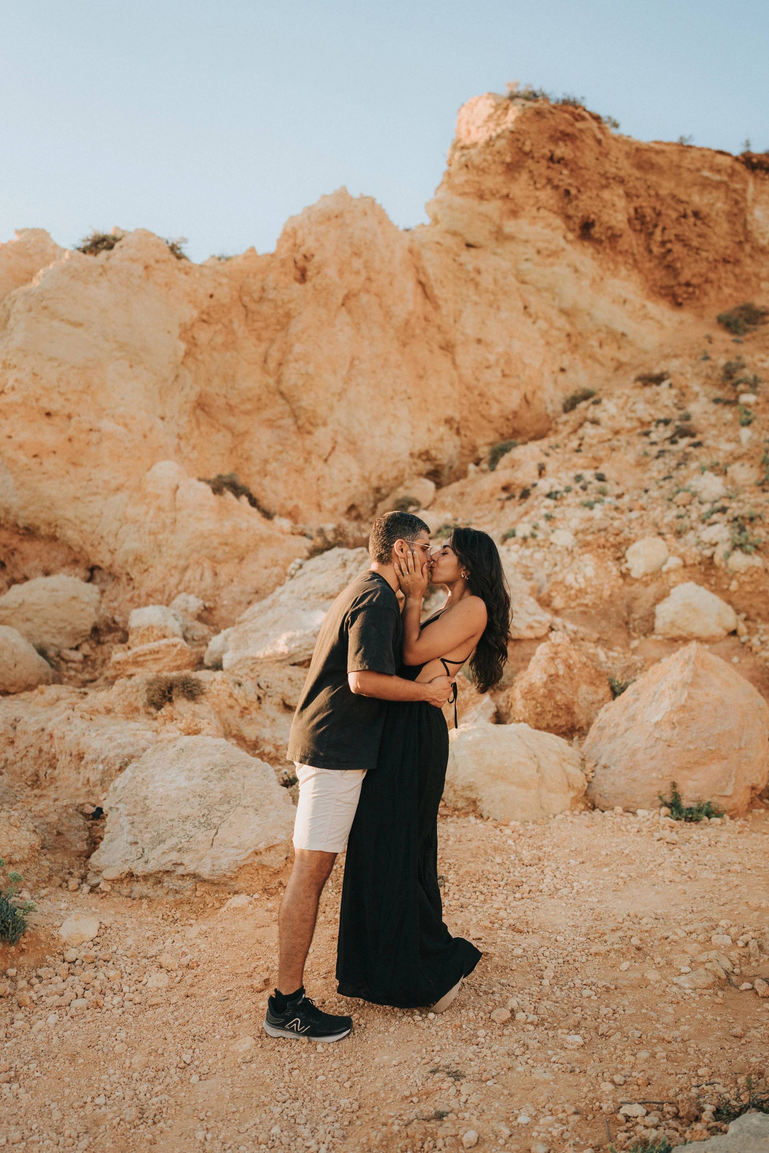 A couple kissing in a rocky desert landscape with large beige rocks and a clear blue sky.