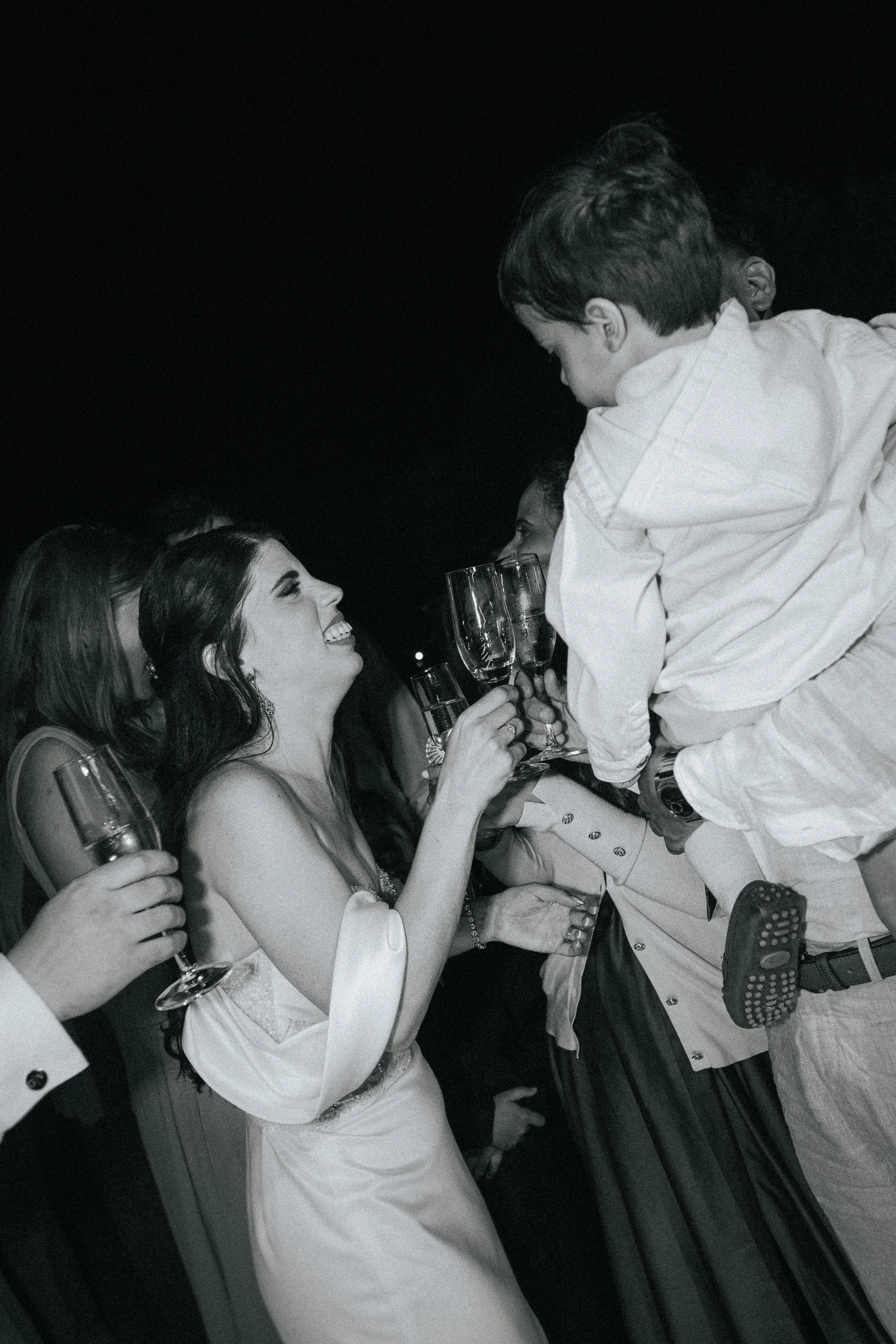 A group of people at a celebration or party, holding drinks, smiling, and engaging in a toast, with a dark background.