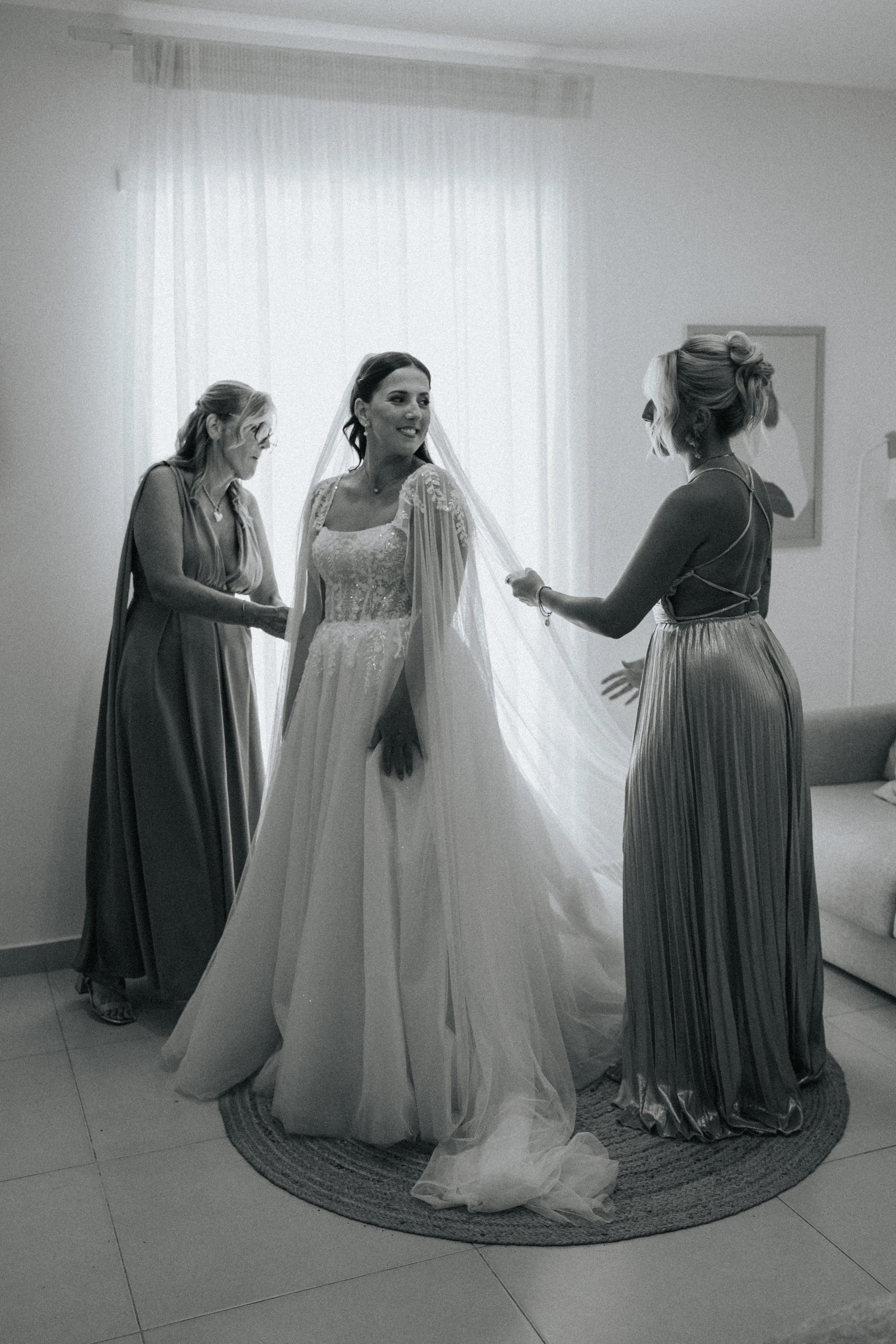 A bride in a wedding dress and veil is getting ready, surrounded by women assisting her, in a room with a curtain and a circular rug on the floor.