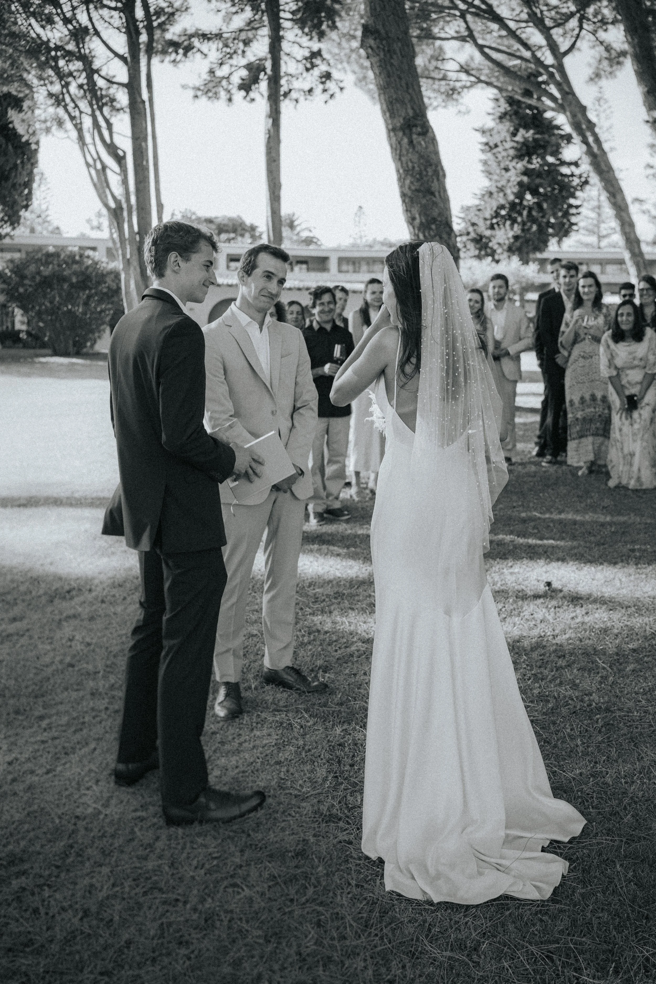 A wedding ceremony taking place outdoors with a bride and groom facing each other, surrounded by guests under tall trees.