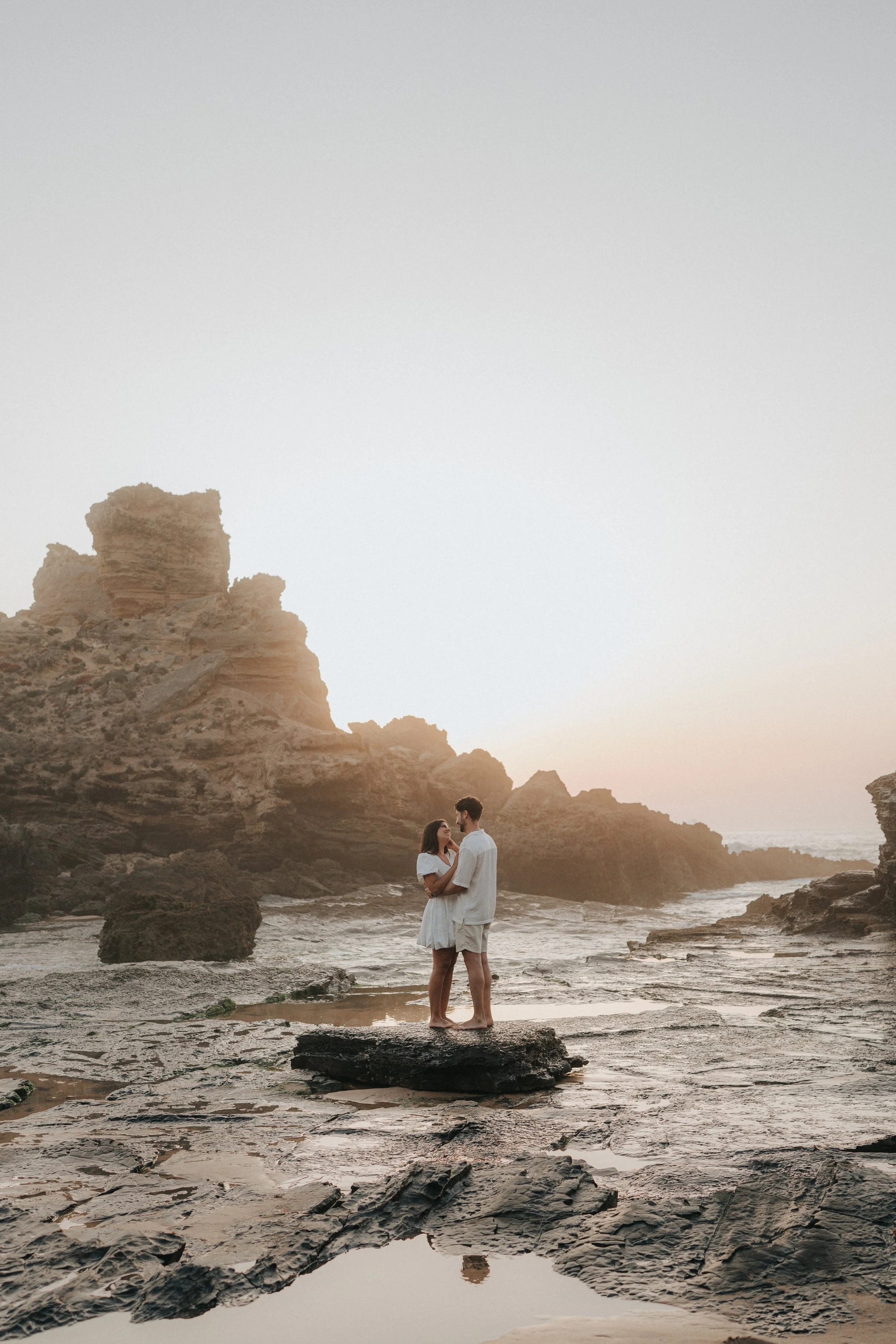 A couple stands on a rocky beach during sunset, holding each other close and gazing into each other's eyes, with large rock formations and calm ocean waves in the background.