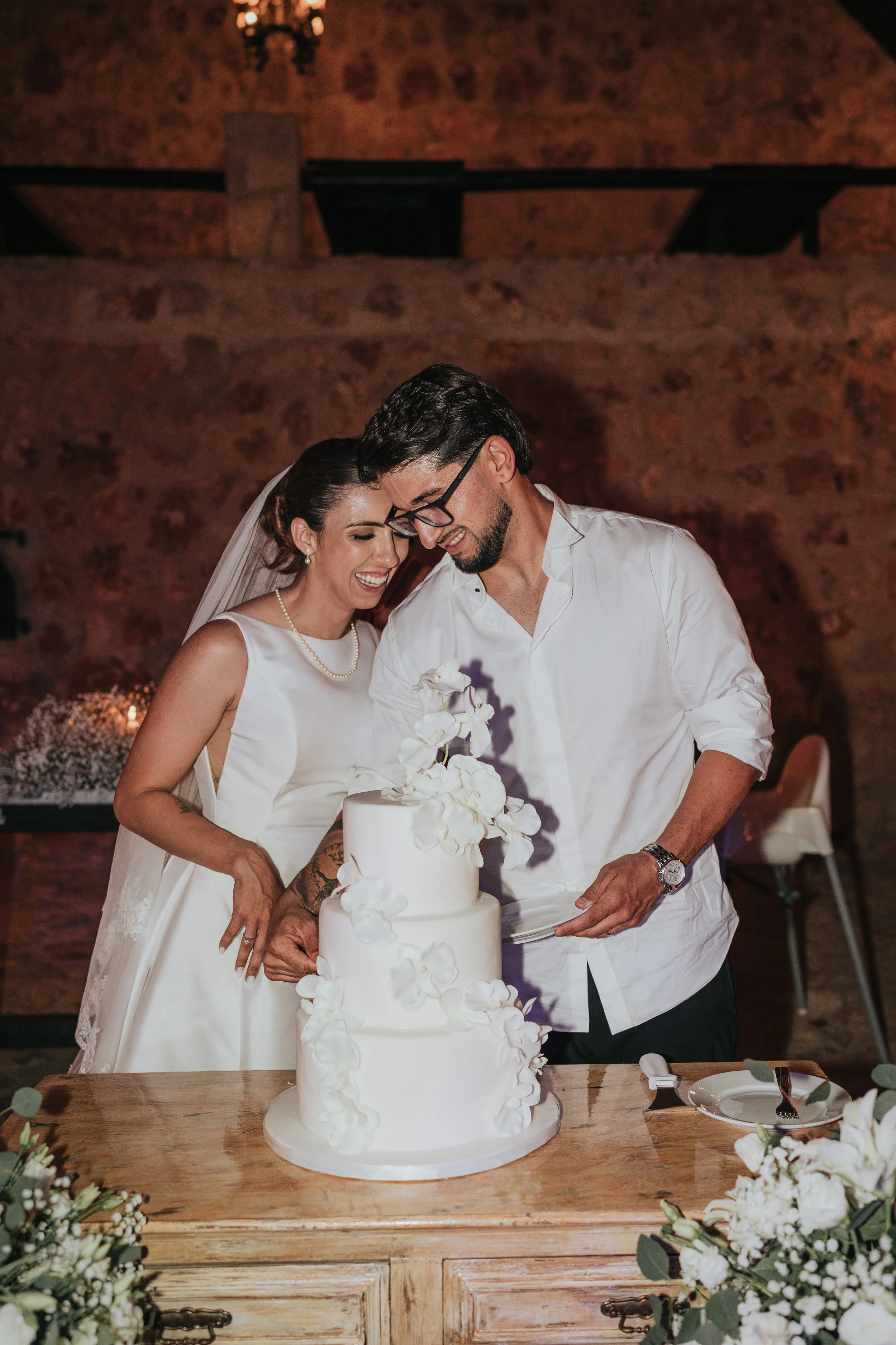 A bride and groom are smiling as they cut a three-tier white wedding cake decorated with white flowers, in a rustic venue with exposed brick walls and floral arrangements on the table.