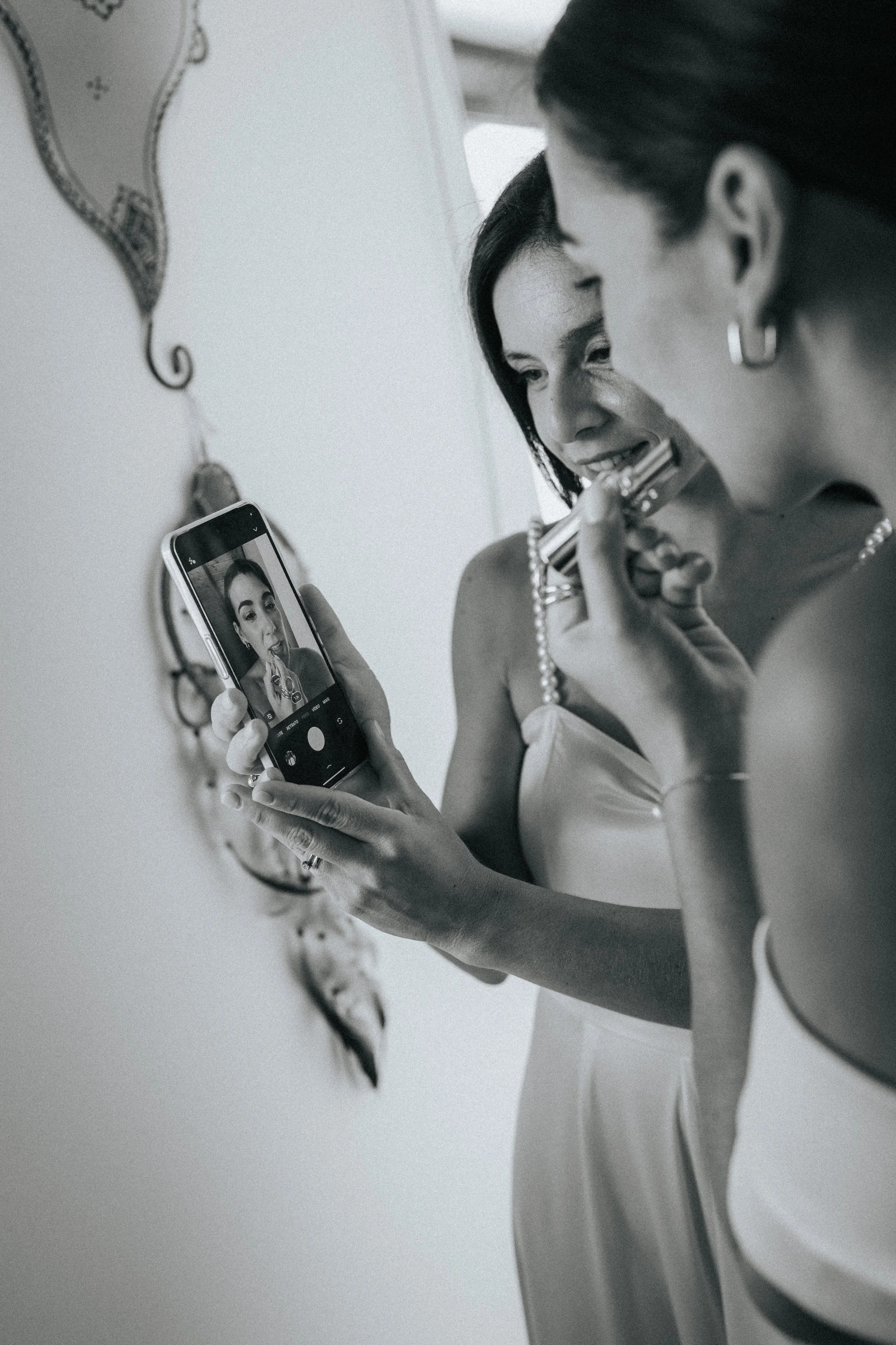 Black and white photo of a woman taking a selfie in front of a mirror, holding her phone with one hand and a lipstick with the other, smiling and looking at her reflection.