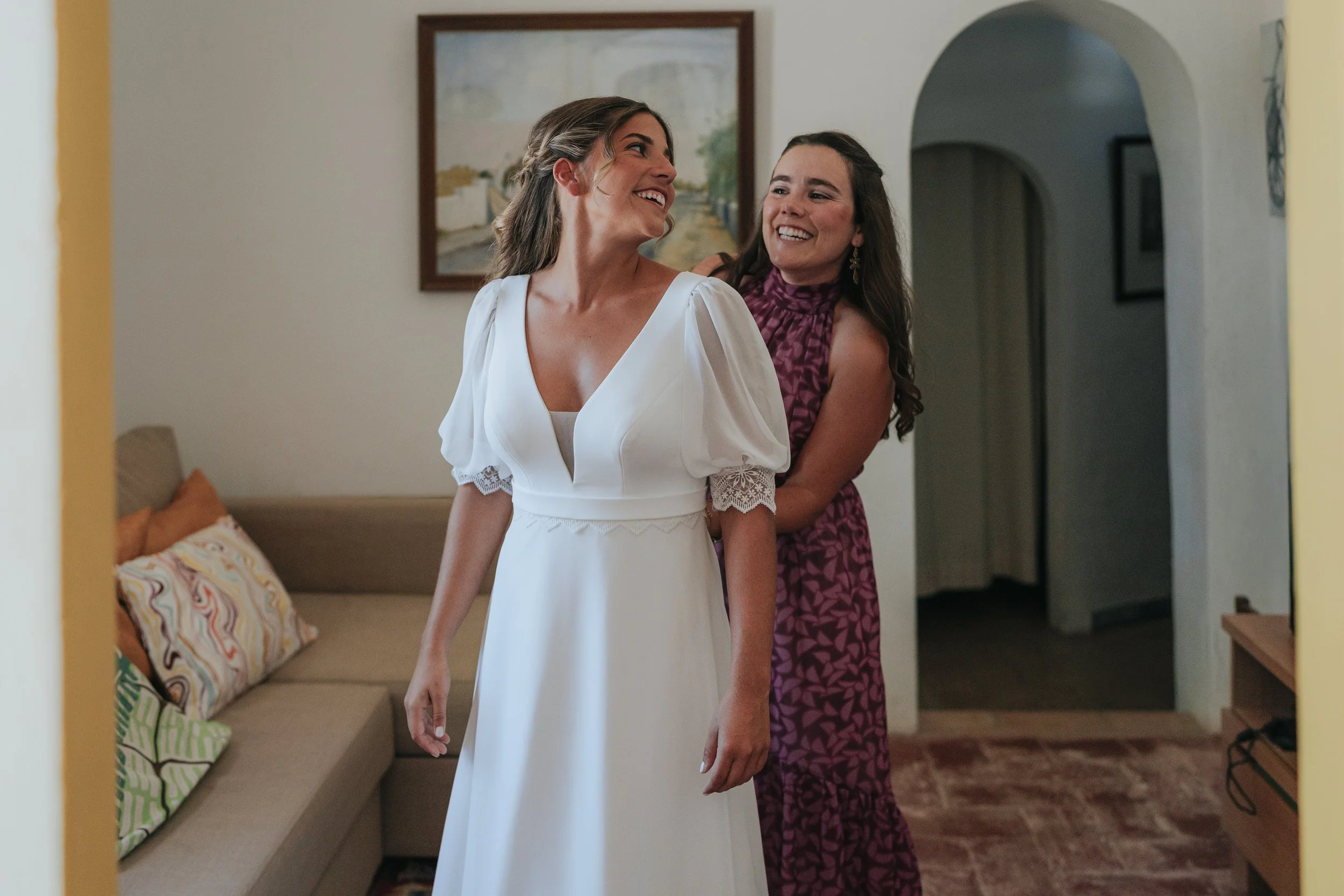 A woman in a white dress is smiling as she is helped into her dress by another woman in a purple patterned dress. They are standing in a living room with a sofa, colorful pillows, and a painting on the wall.
