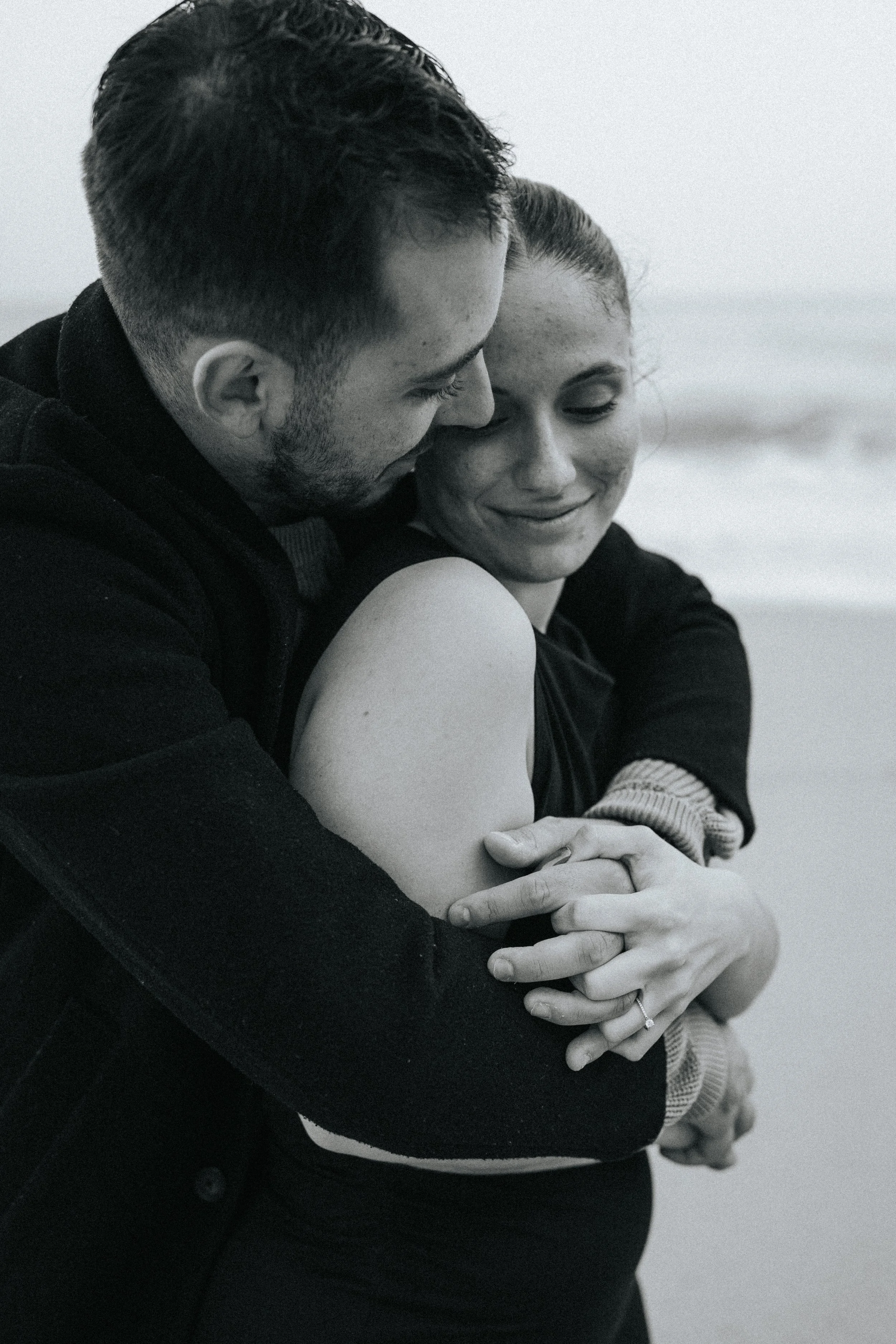 A black and white photo of a couple embracing each other, with the man gently kissing the woman's forehead and both smiling softly, at the beach.