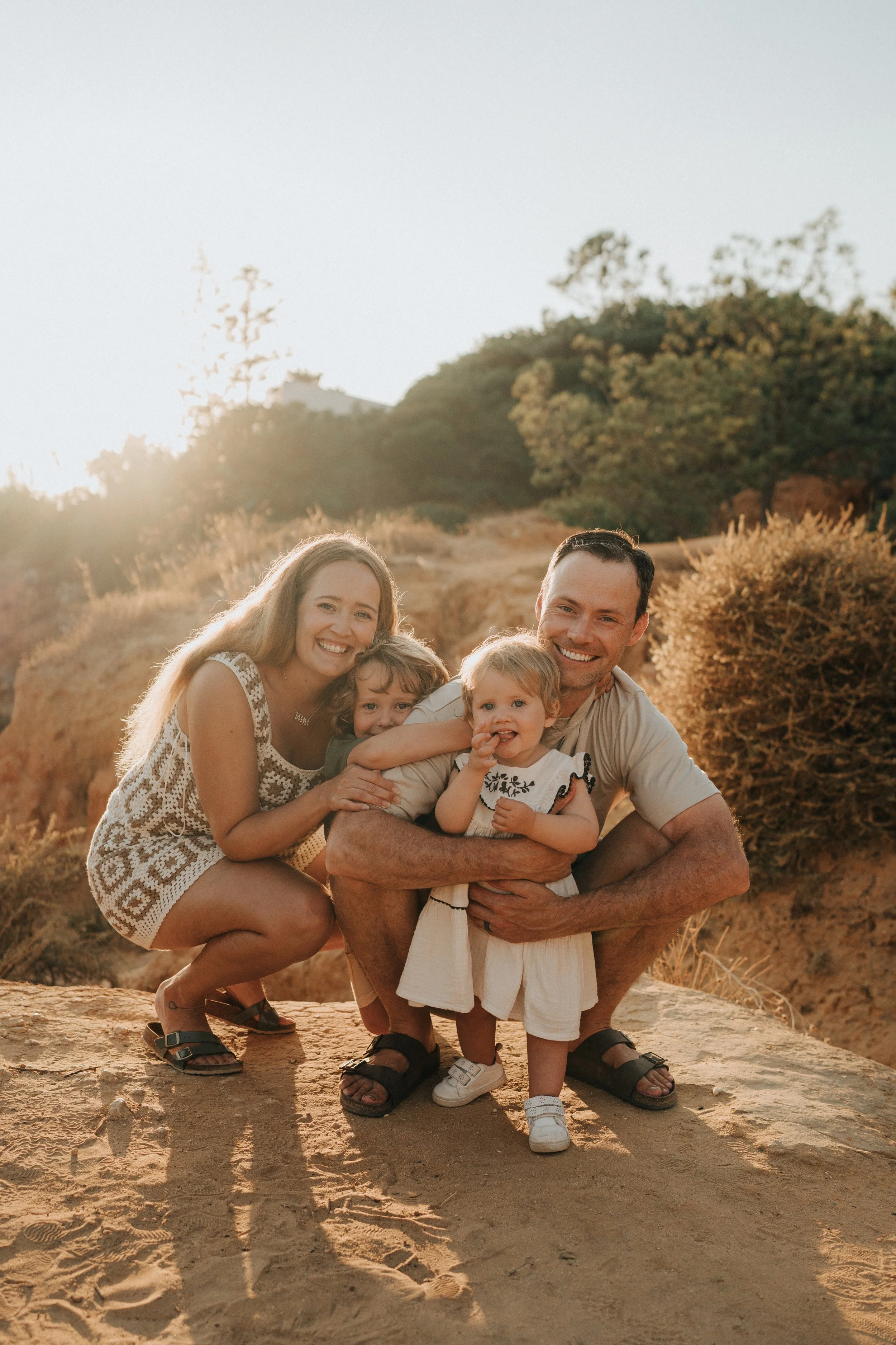 A happy family of five with two young children outdoors on a sunny day, surrounded by trees and dry terrain, smiling and posing closely together.