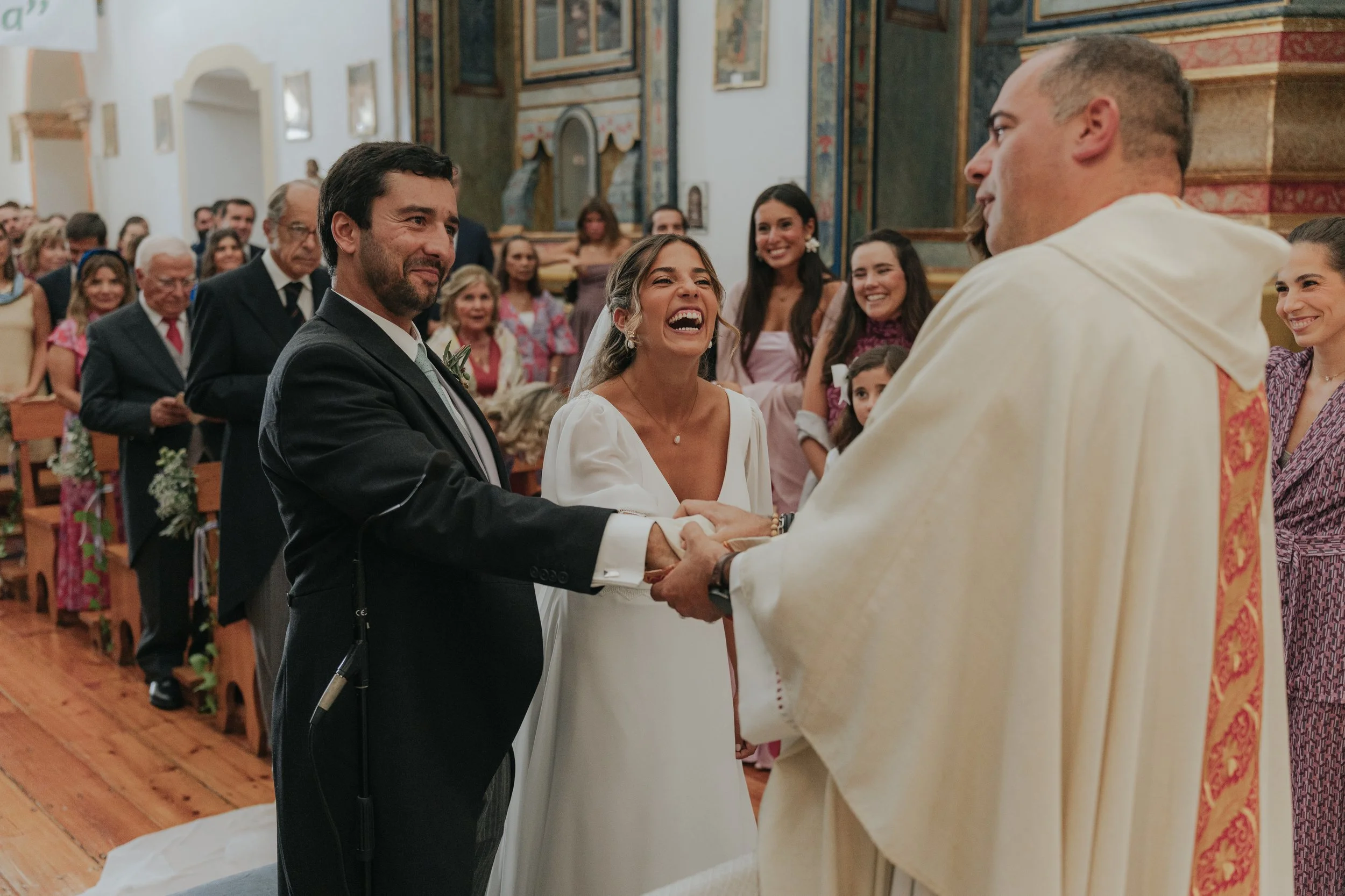 A bride and groom holding hands and smiling during their wedding ceremony inside a church, with guests smiling in the background.