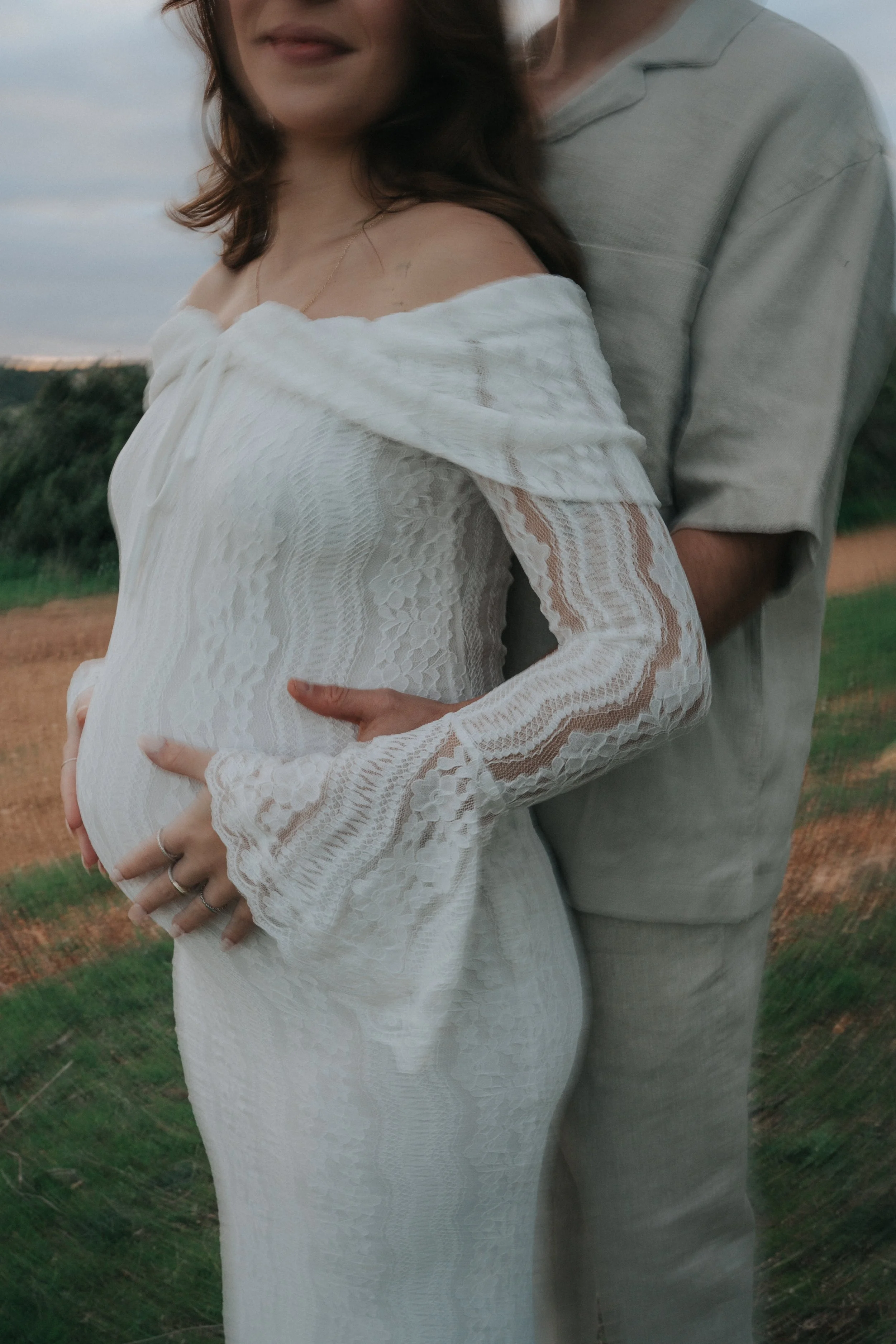 A pregnant woman in a white lace dress is standing outdoors with a man behind her, both holding her belly. The woman has shoulder-length brown hair and is wearing a delicate necklace. The man is wearing a light gray shirt. The background features a f