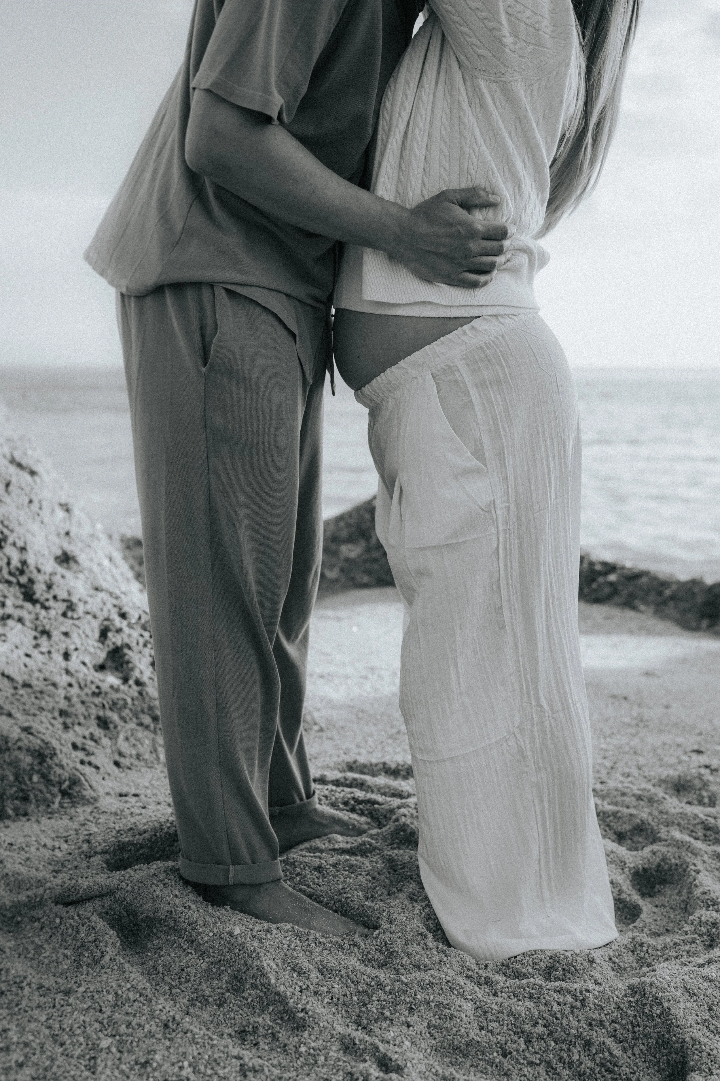 A couple standing on a sandy beach, holding each other closely. The man is dressed in casual gray pants and a t-shirt, while the woman is wearing light-colored, loose-fitting pants and a long-sleeve top. The image is in black and white, capturing an 