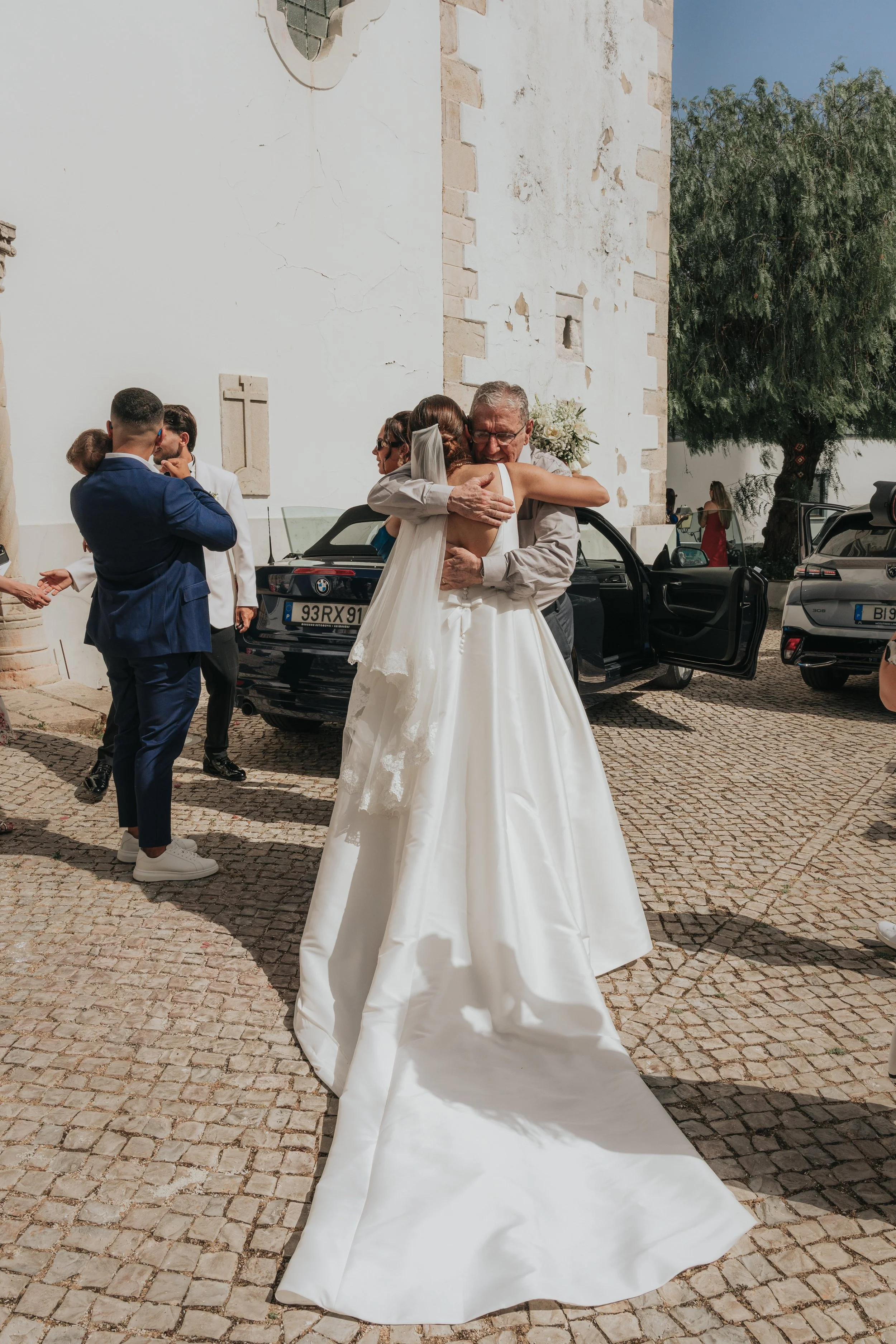 A bride and an elderly man sharing a hug at a wedding ceremony outdoors, with guests and cars in the background.