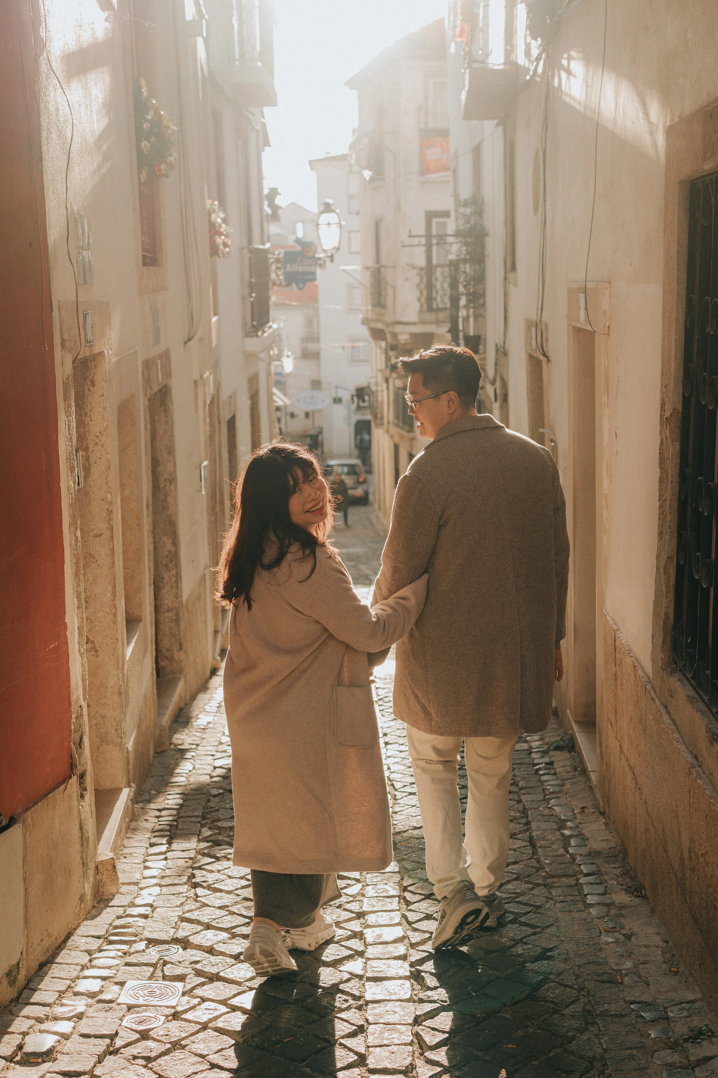 A couple walking hand-in-hand down a cobblestone alleyway in a European city during the daytime, smiling and looking at each other, with old buildings and a pastel-colored wall on the side.