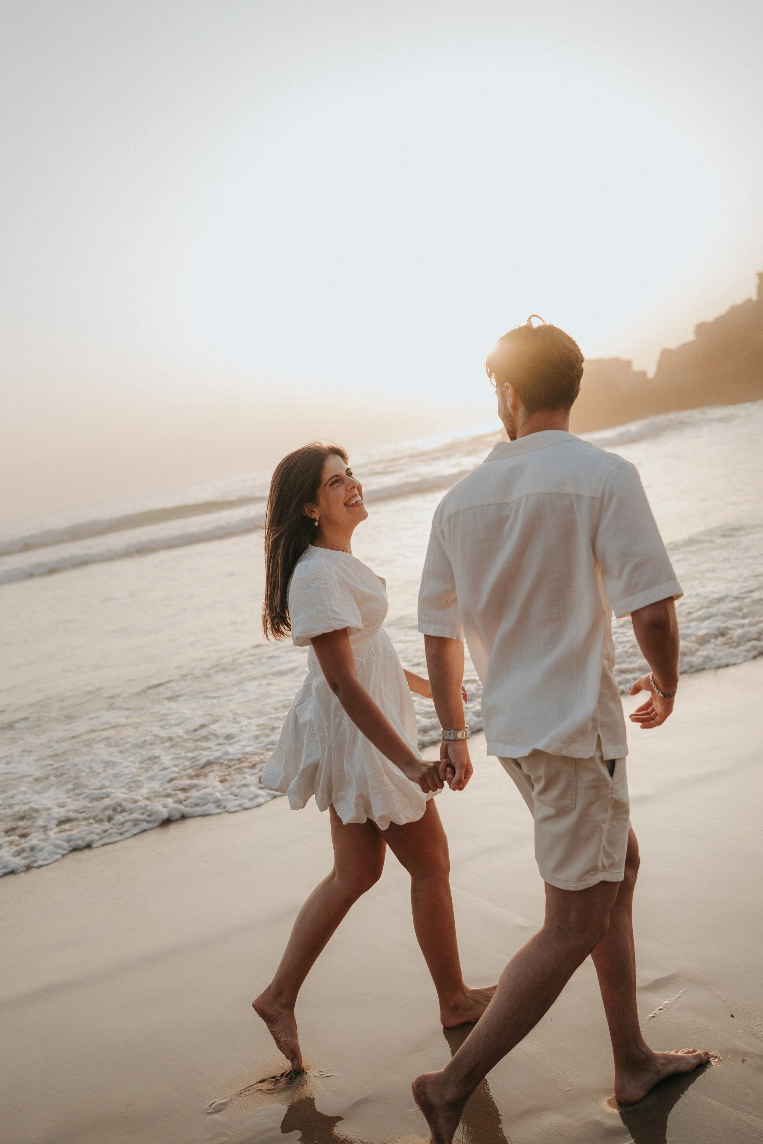 A couple holding hands and walking on the beach at sunset, smiling and enjoying each other's company.