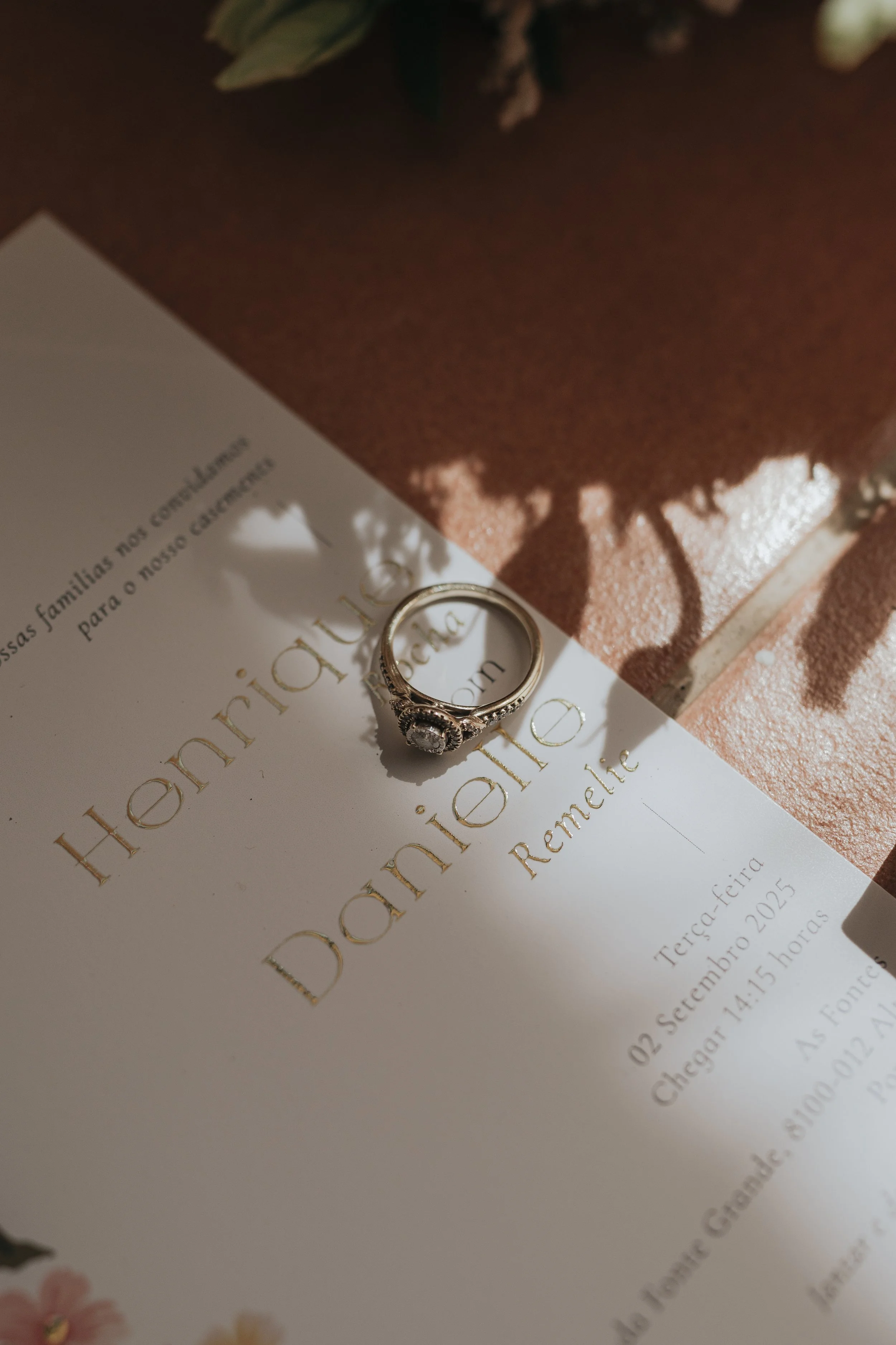A wedding invitation for a couple named Thalita and Daniel, with gold embossed text, partially visible. A silver engagement ring is placed on the invitation. In the background, there is a brown textured gift box and some floral decorations.