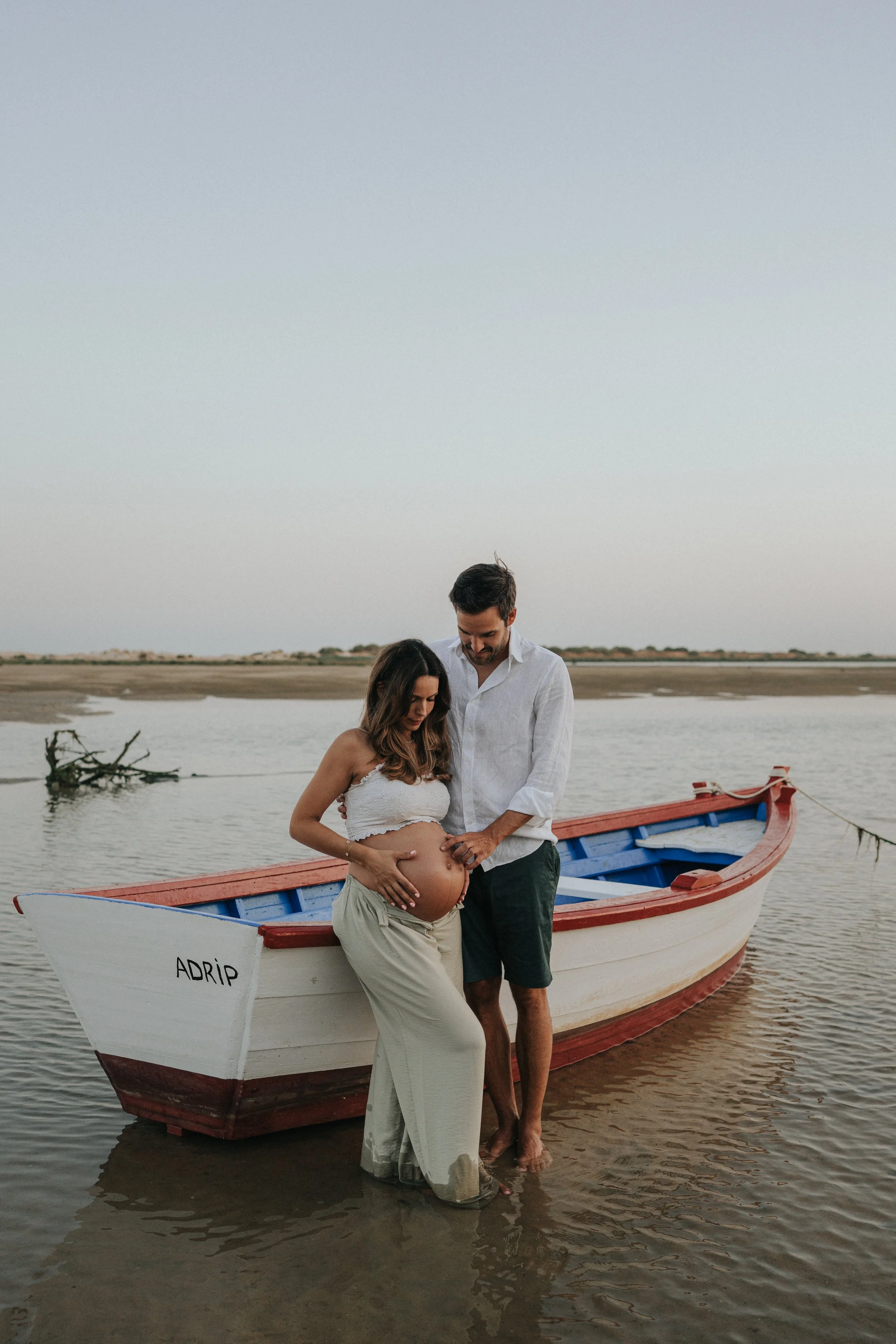 A pregnant woman in beige pants and a white cropped top, and a man in a white shirt and dark shorts, standing barefoot in shallow water near a boat named 'ADRIP' on a calm river or lake at sunset or dusk.