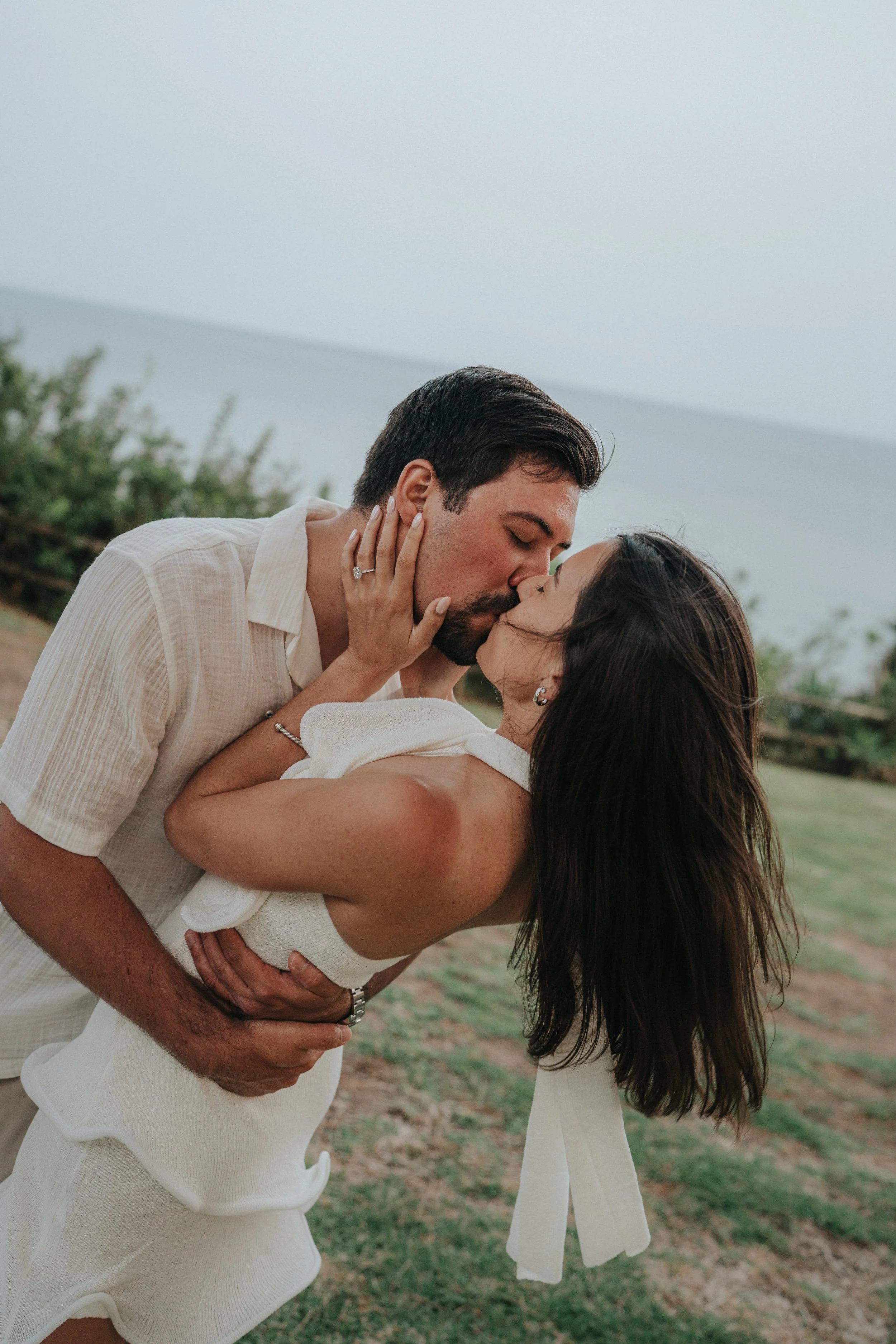 A man and woman in a loving embrace, sharing a kiss outdoors near a grassy area with a body of water and mountains in the background.