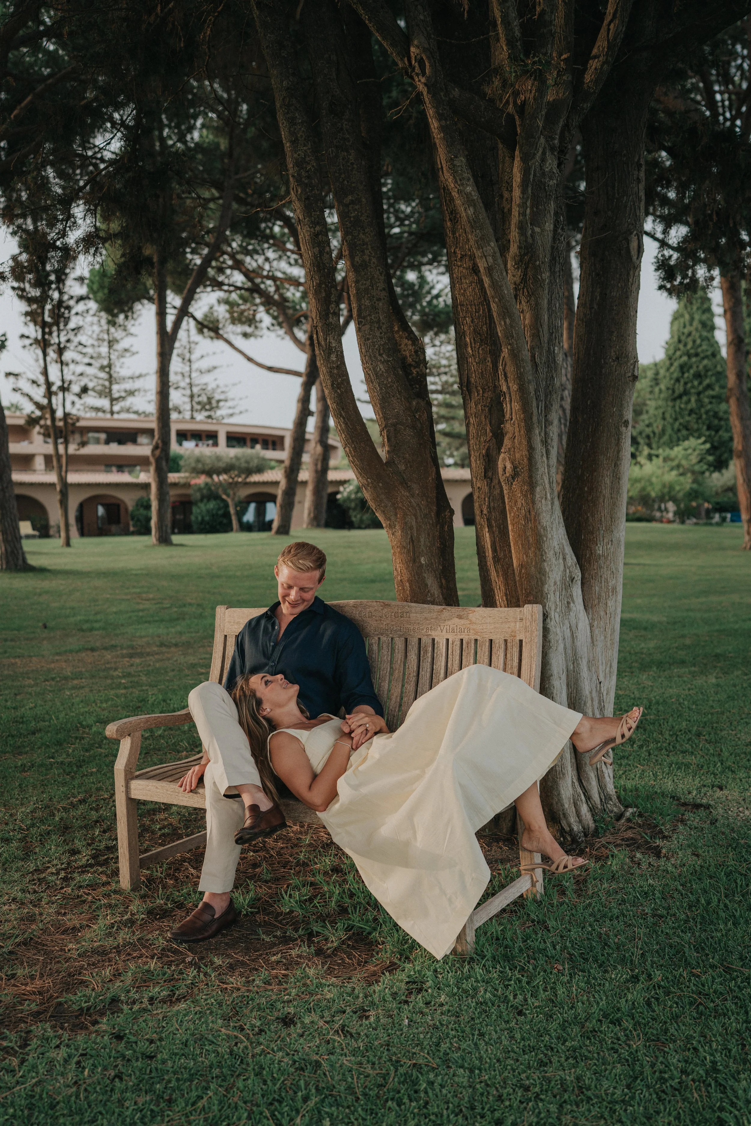 A young man and woman sitting and lying on a wooden park bench under a tree. The woman is lying with her head in the man's lap, smiling and looking up at him, while he looks down at her with a happy expression. The woman is wearing a yellow dress and