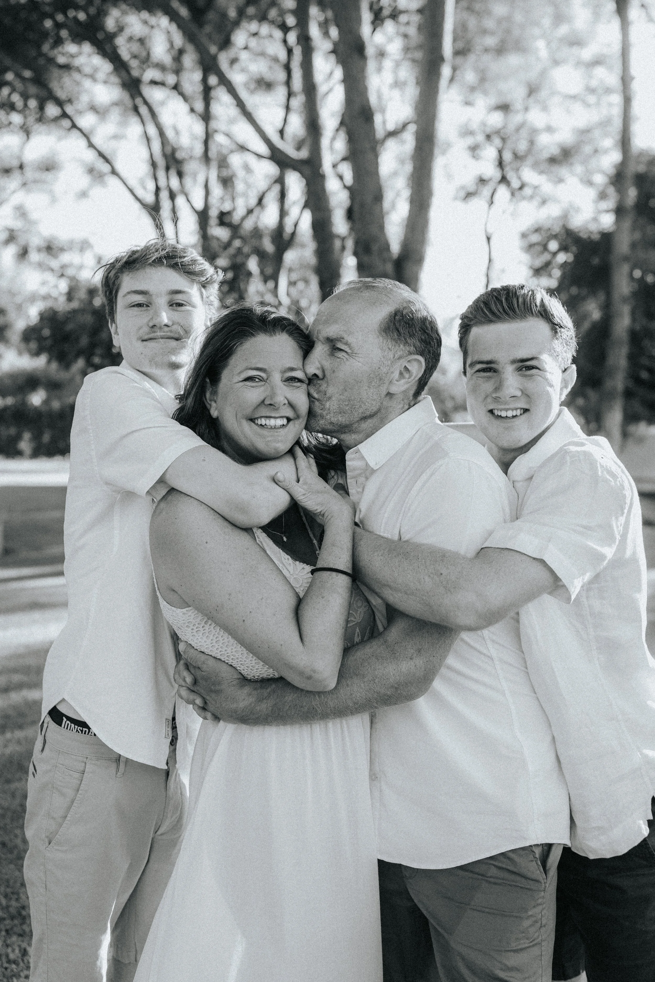 A group of five people, including an older woman and man, showing affection with hugs and kisses outdoors, with trees in the background.