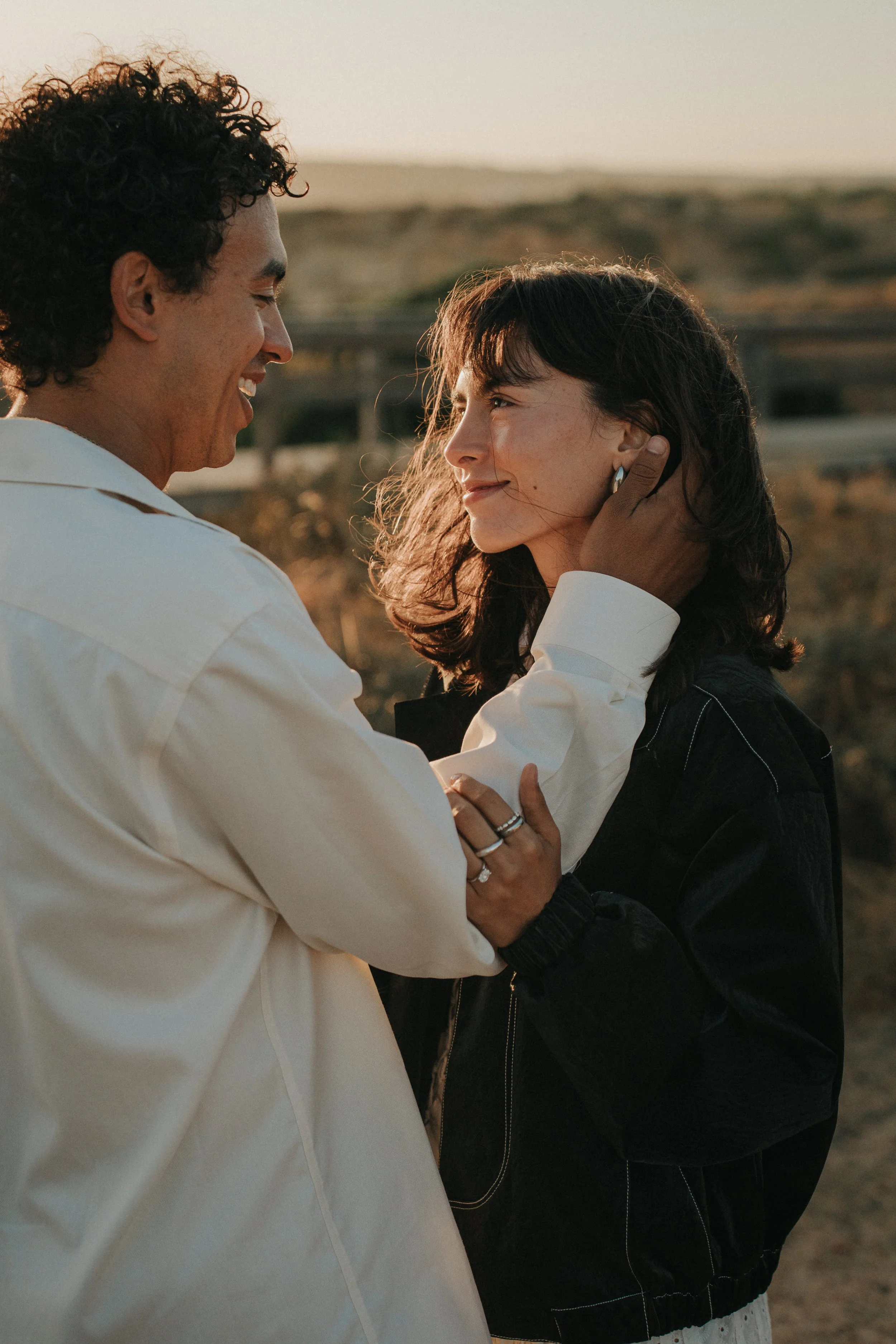 A man and woman face to face, sharing an affectionate moment outdoors during sunset. The man holds the woman's face gently with his hand, smiling warmly. The woman gazes back with a soft smile, her hand resting lightly on the man's arm.