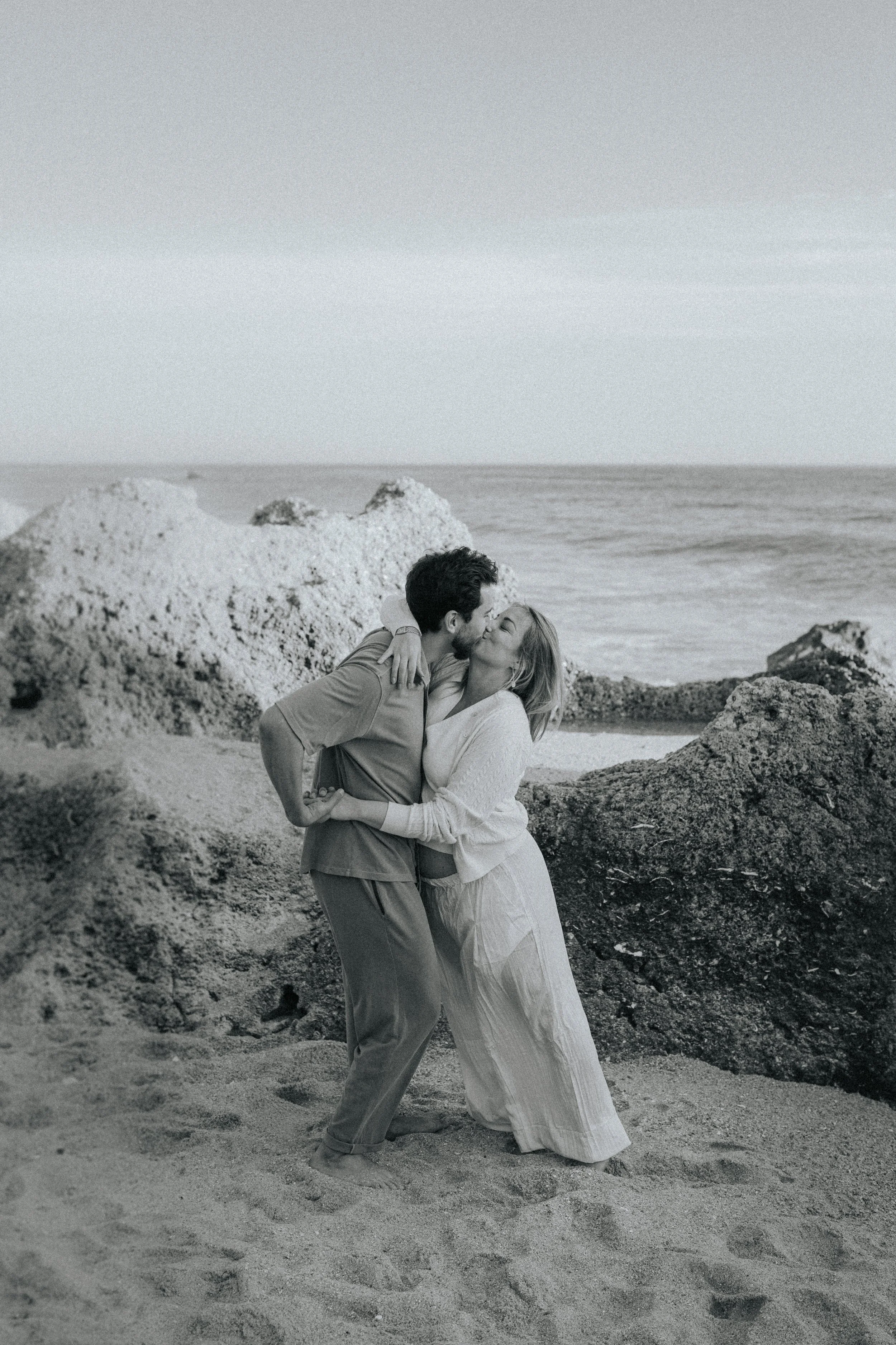 A black and white photo of a couple kissing on a rocky beach, surrounded by rocks and sand, with the ocean and sky in the background.