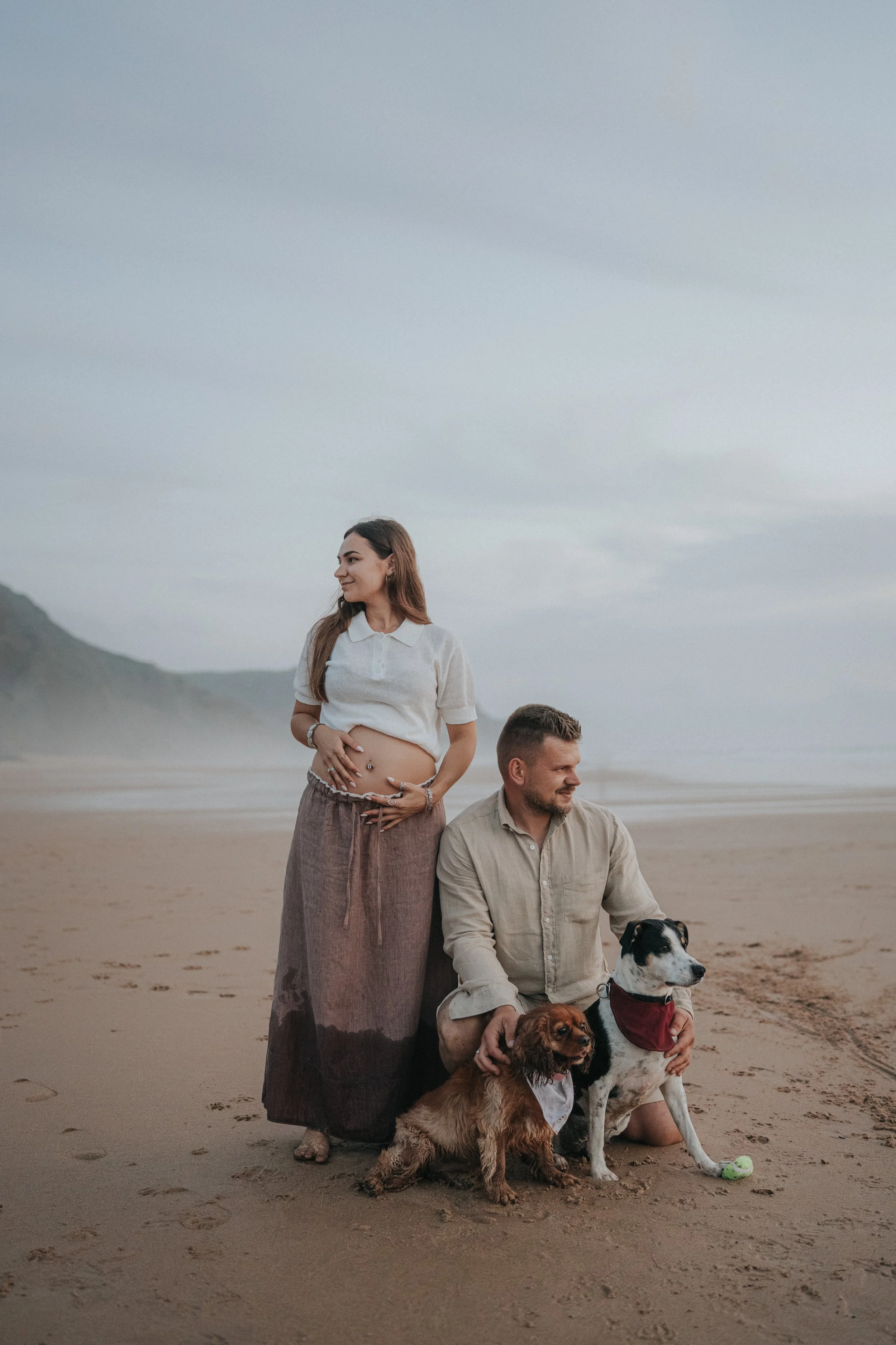 A woman standing on a beach with a pregnant belly, wearing a white top and long skirt, looking to her left. A man kneeling on the sand with two dogs, one sitting and one lying down, holding a tennis ball. The background features dunes and a cloudy sk