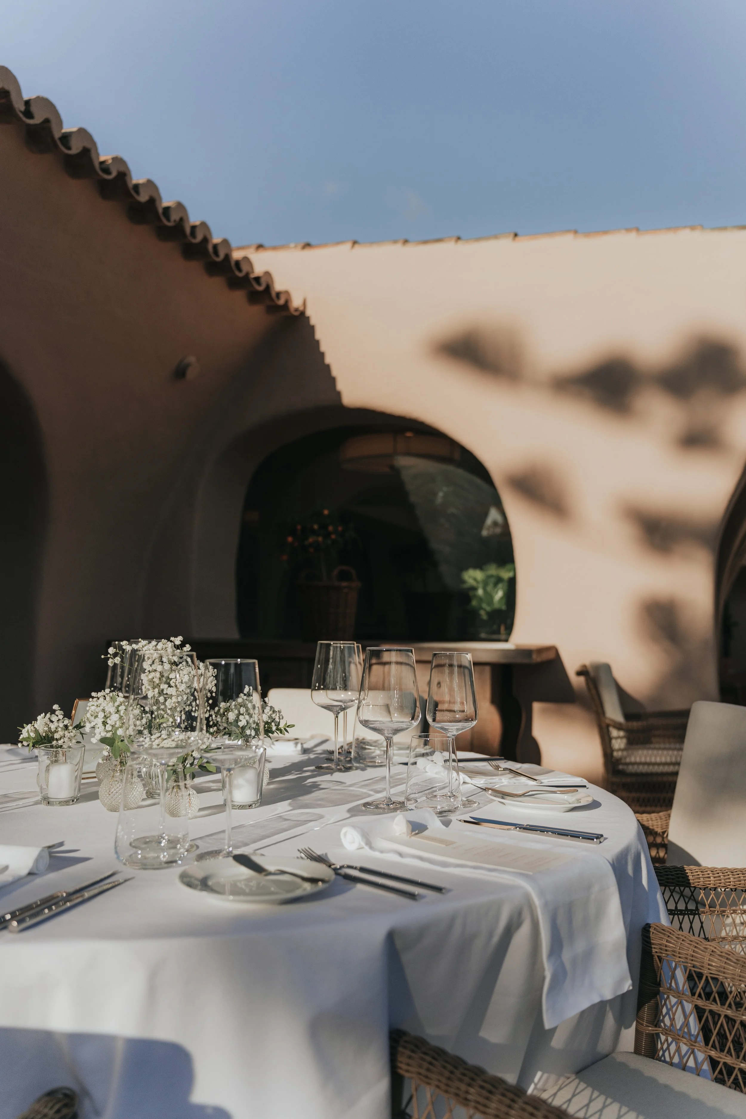 Elegant outdoor dining table set with white tablecloth, floral centerpiece, wine glasses, and silverware, under a shaded patio with ceiling beams and a clear blue sky.