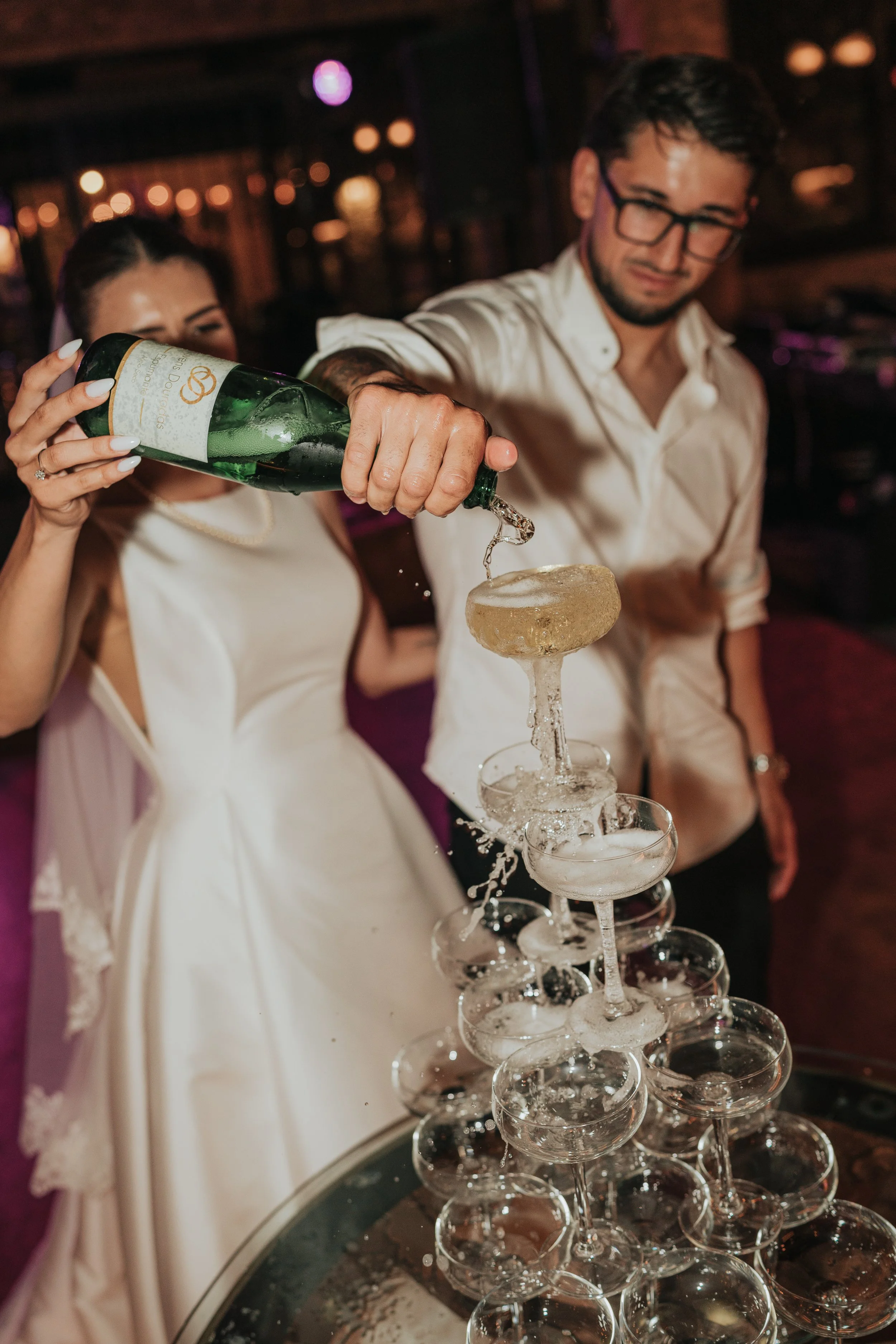 A couple standing at a wedding reception, pouring champagne into a tower of glasses.
