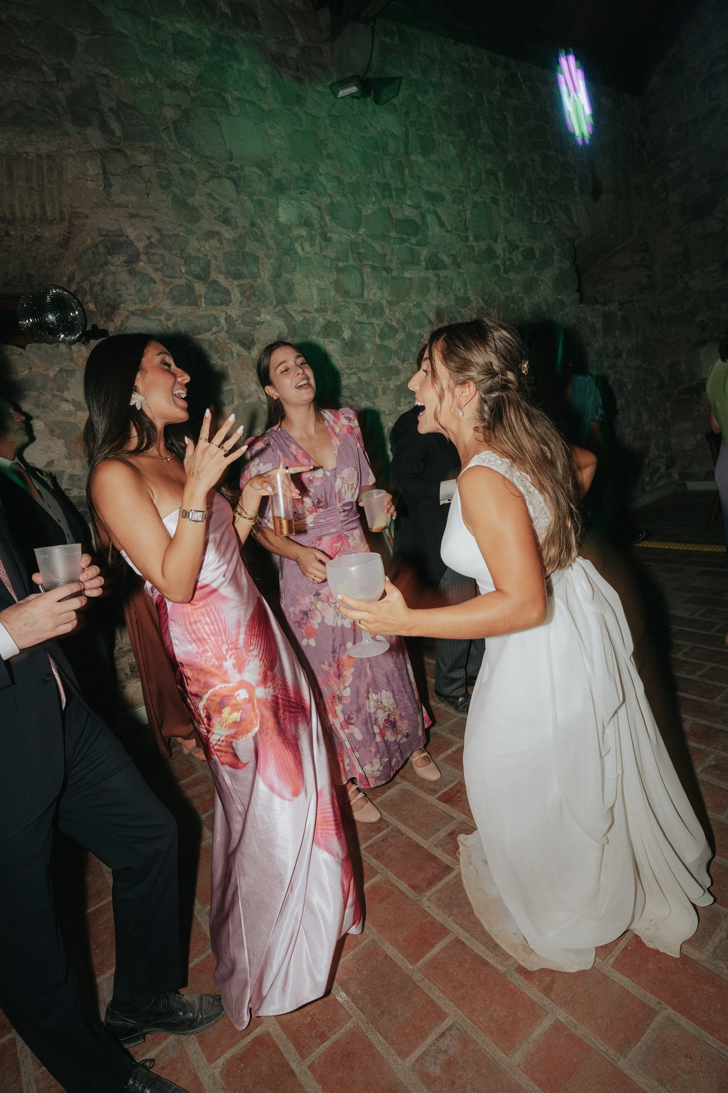 Women in formal dresses celebrating and laughing at a party with drinks in hand, in a rustic setting with stone walls and a neon light.