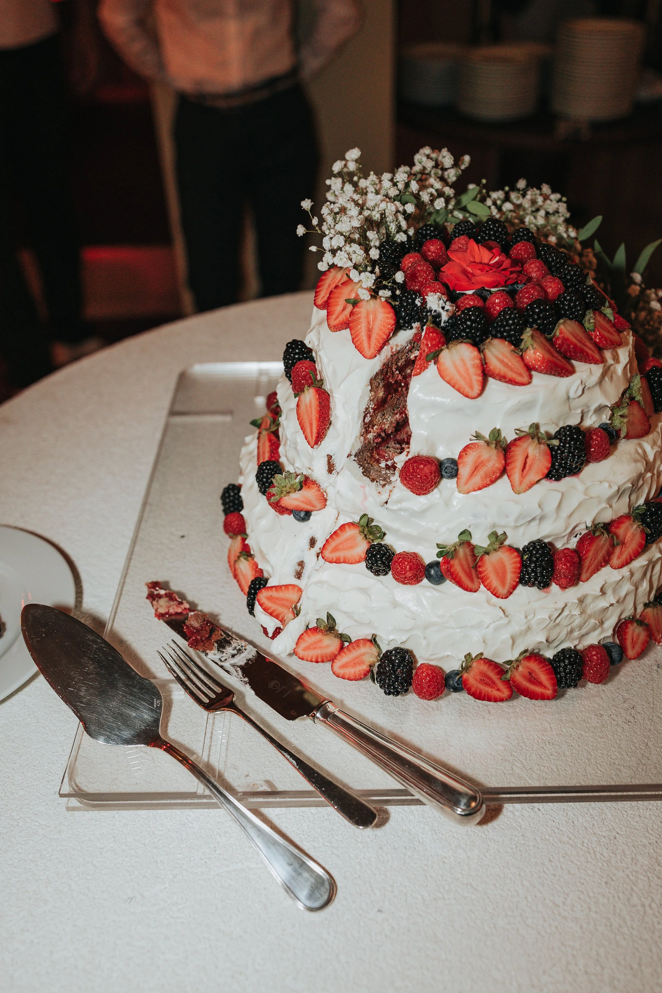 A layered white frosted cake decorated with sliced strawberries, blackberries, raspberries, and white baby's breath flowers, with a slice cut out and a cake knife and server placed on a tray in front.