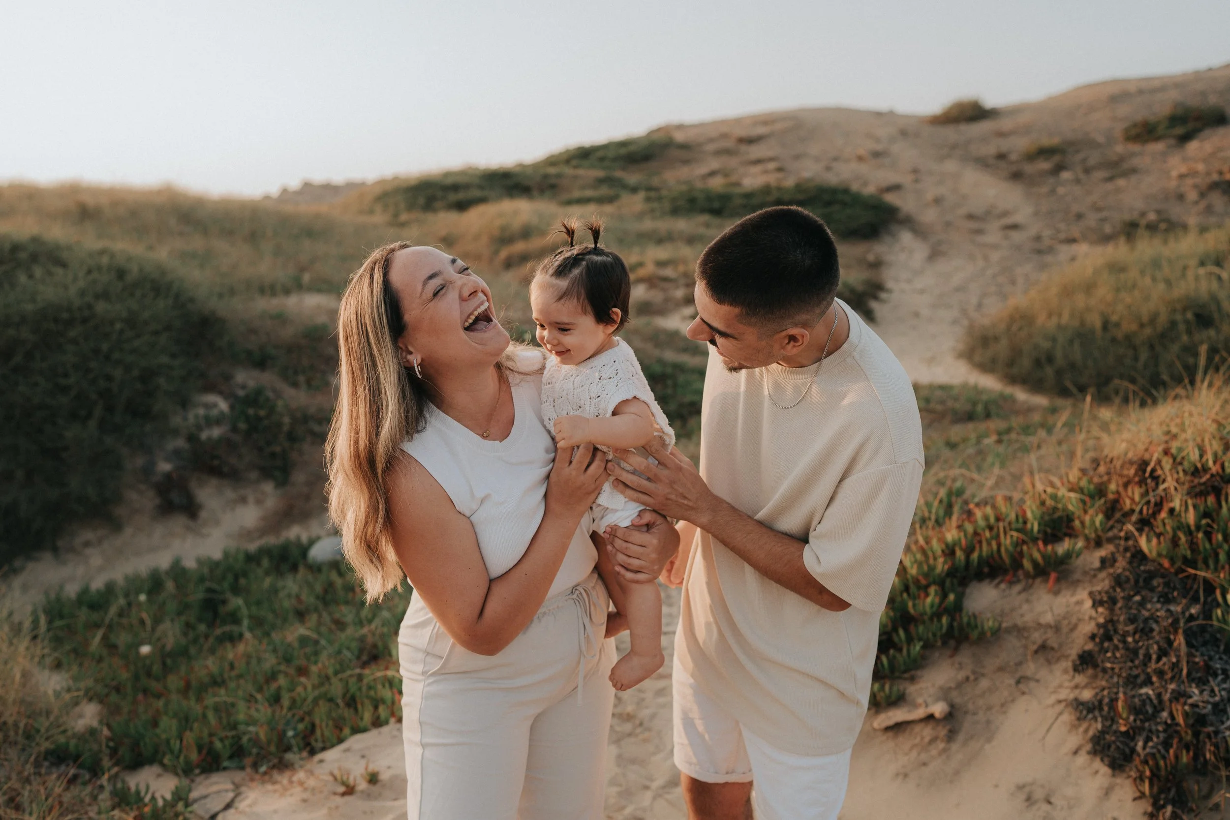 A happy woman holding a young girl, standing on a beach with rolling hills in the background. The woman and girl are smiling and laughing. A young man is standing beside them, looking at the girl with a smile, all dressed in light-colored clothing.