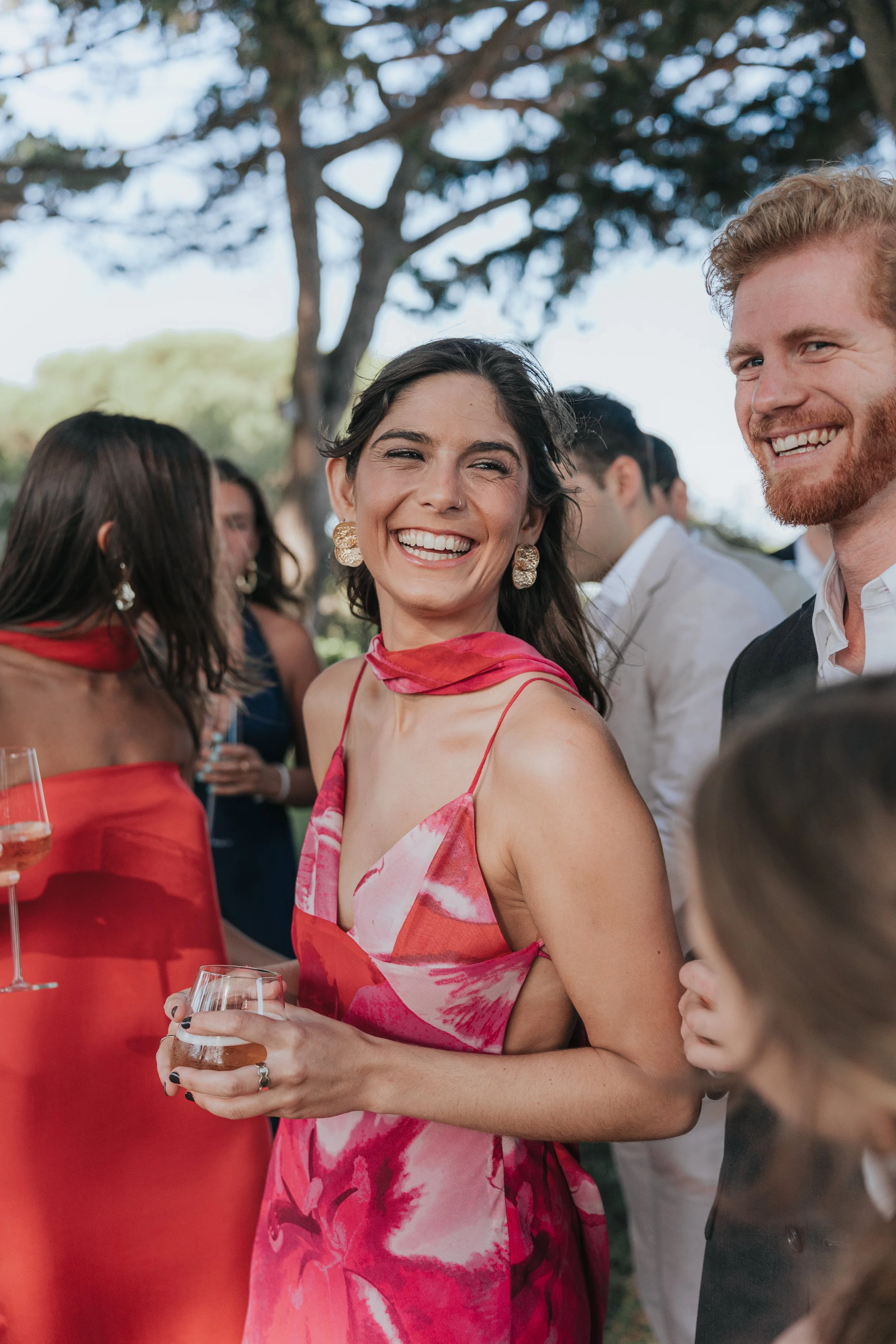 A woman in a pink dress smiling at an outdoor social gathering, holding a glass of wine, with other people dressed formally in the background.