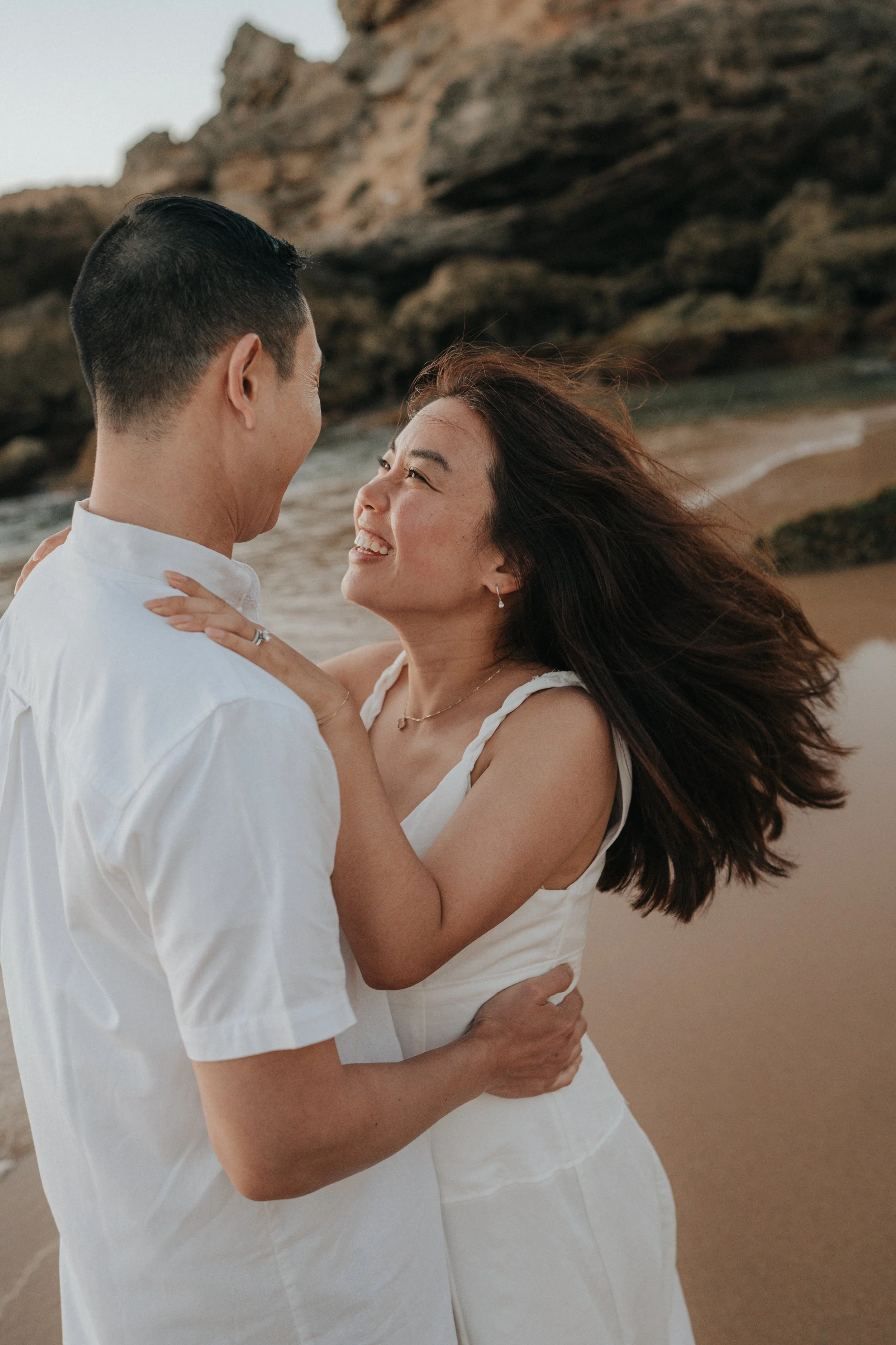 A couple dancing on the beach with rocks in the background, smiling and looking at each other.