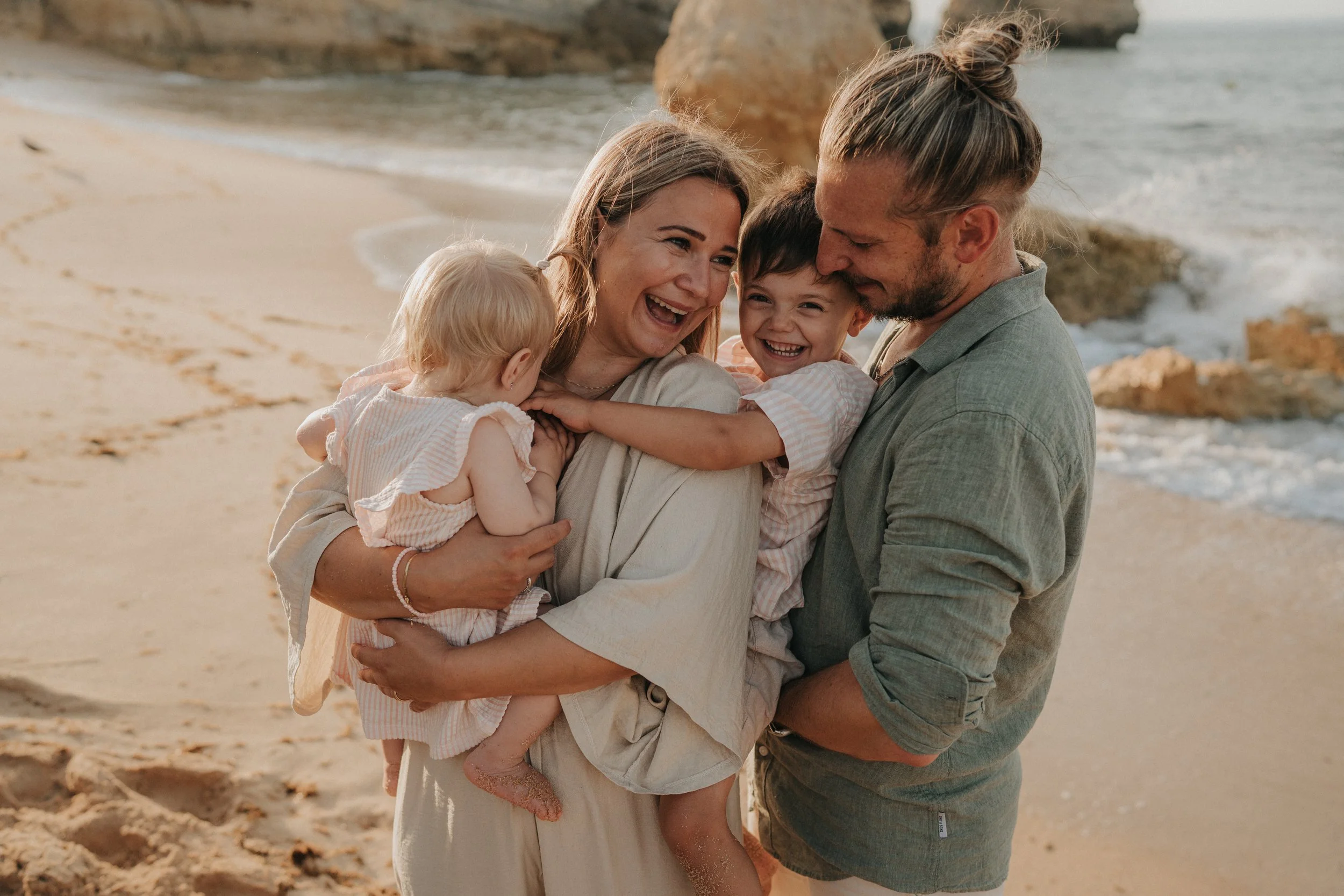 A happy family of four embracing each other on a sandy beach, with rocky formations and the ocean in the background.