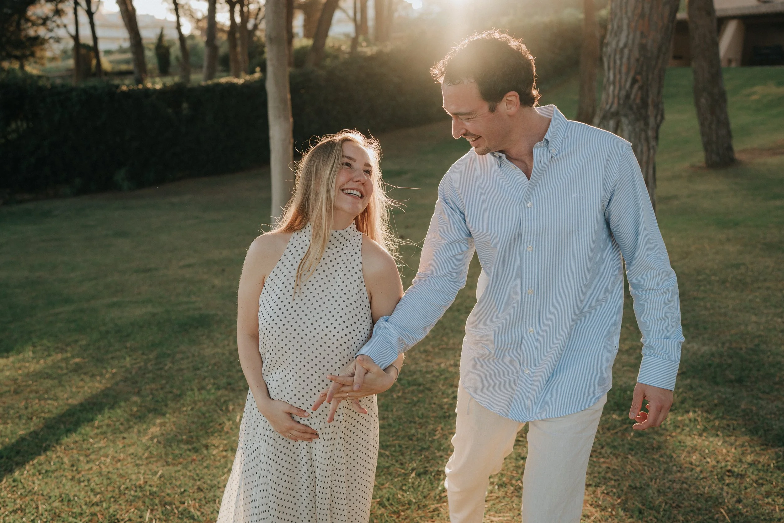 A couple holding hands and smiling outdoors in a park during sunset, with trees in the background.