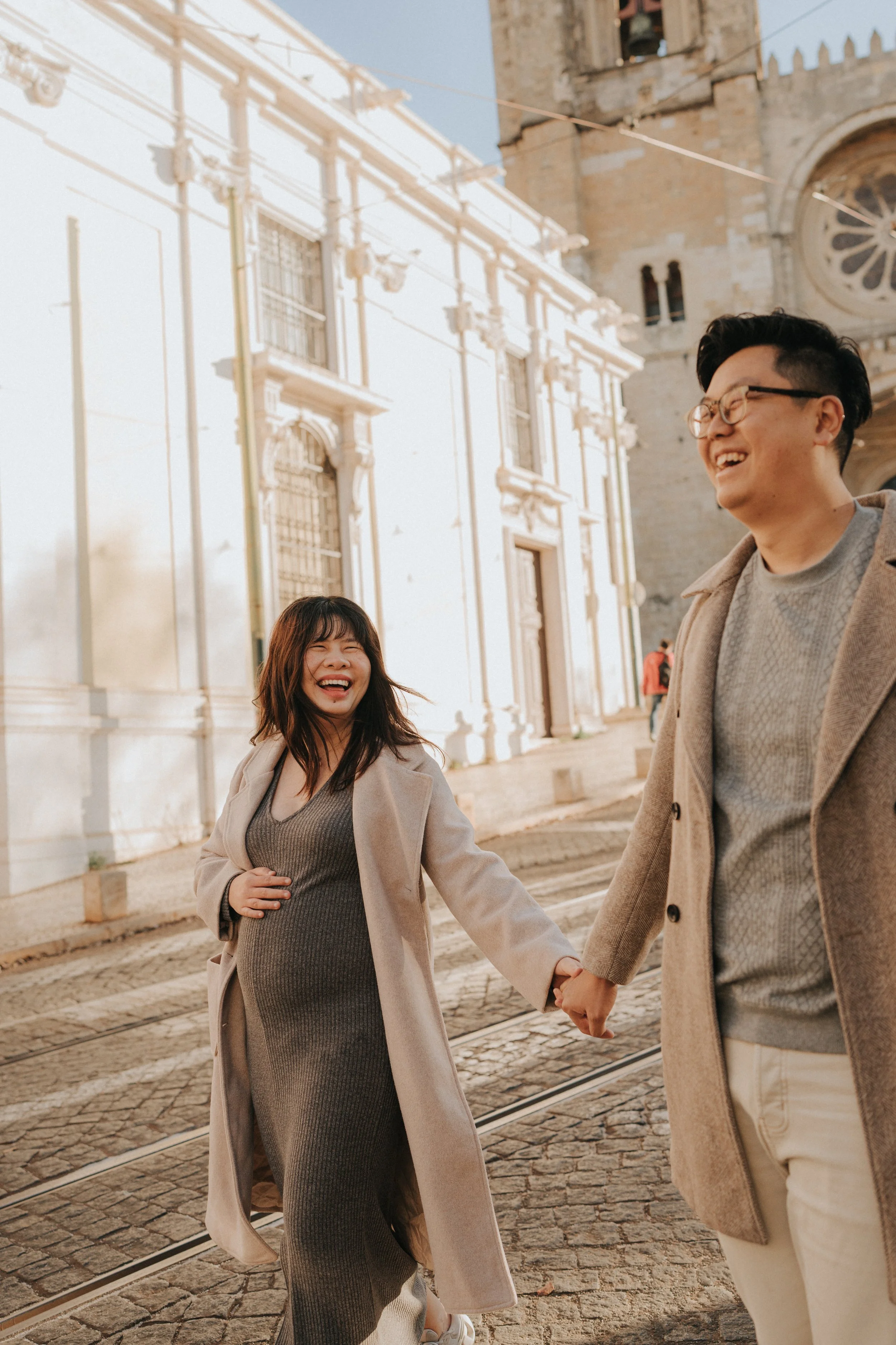 Two smiling people, one woman with dark hair and a man with dark hair and glasses, holding hands and walking outdoors in front of historic buildings.