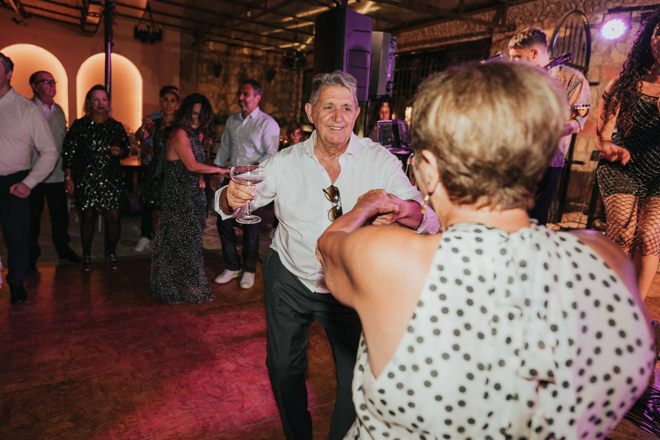 Older man dancing with woman wearing a polka-dot shirt at a lively party or celebration in a rustic venue with stone walls and colorful lighting.