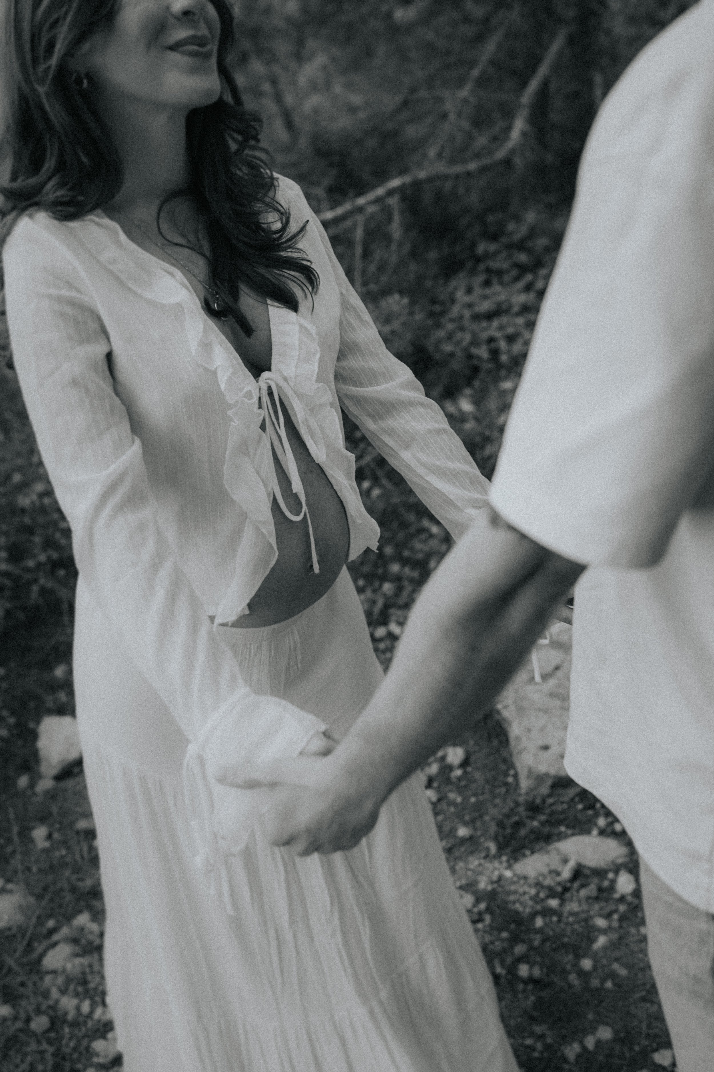 A woman with wavy hair smiling, wearing a flowing white dress with a tie-front top, holding hands with another person outdoors.