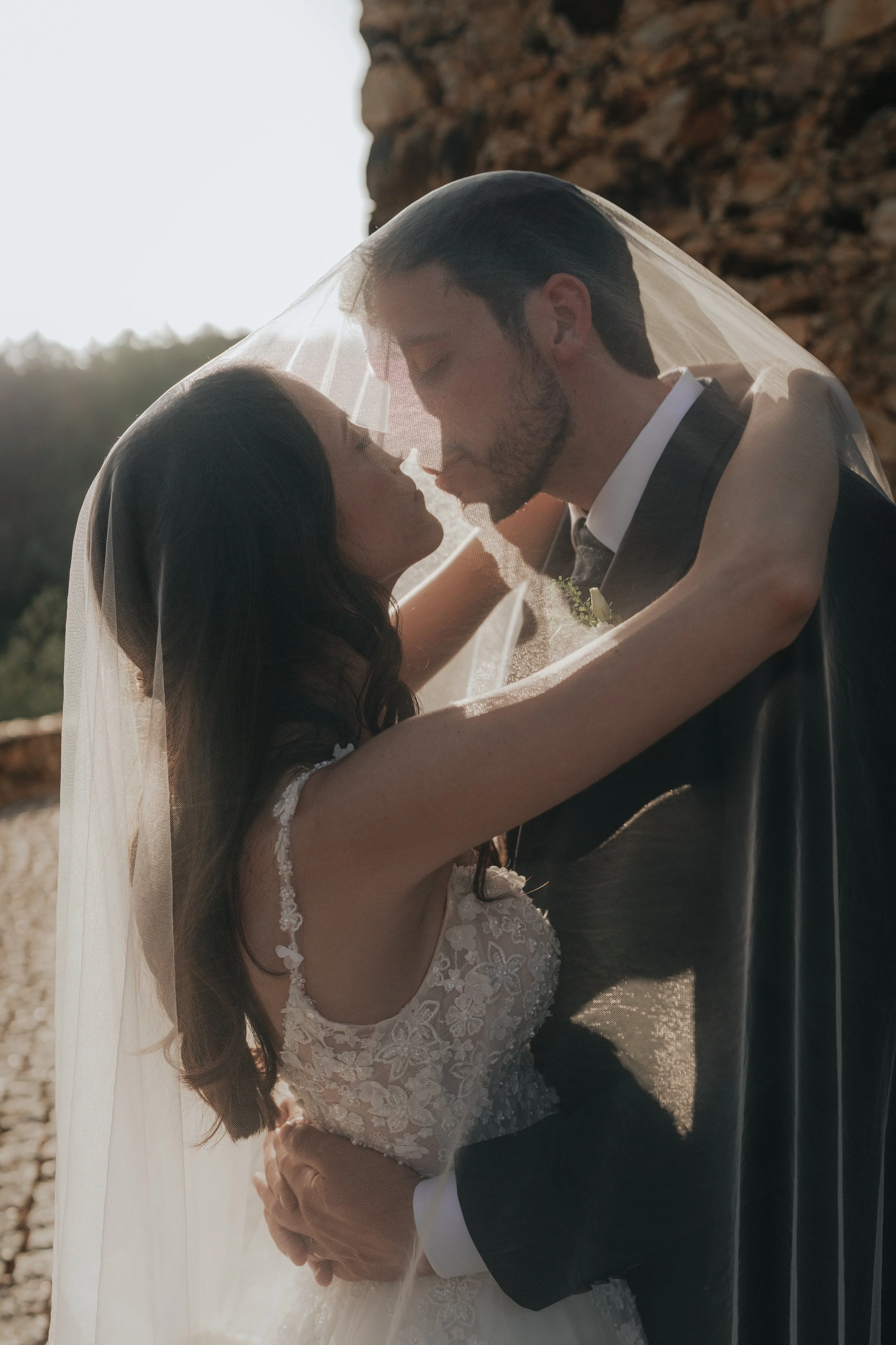 A bride and groom are hugging and about to kiss, with the bride wearing a lace wedding dress and a veil, and the groom in a tuxedo, outdoors near a stone wall during sunset.