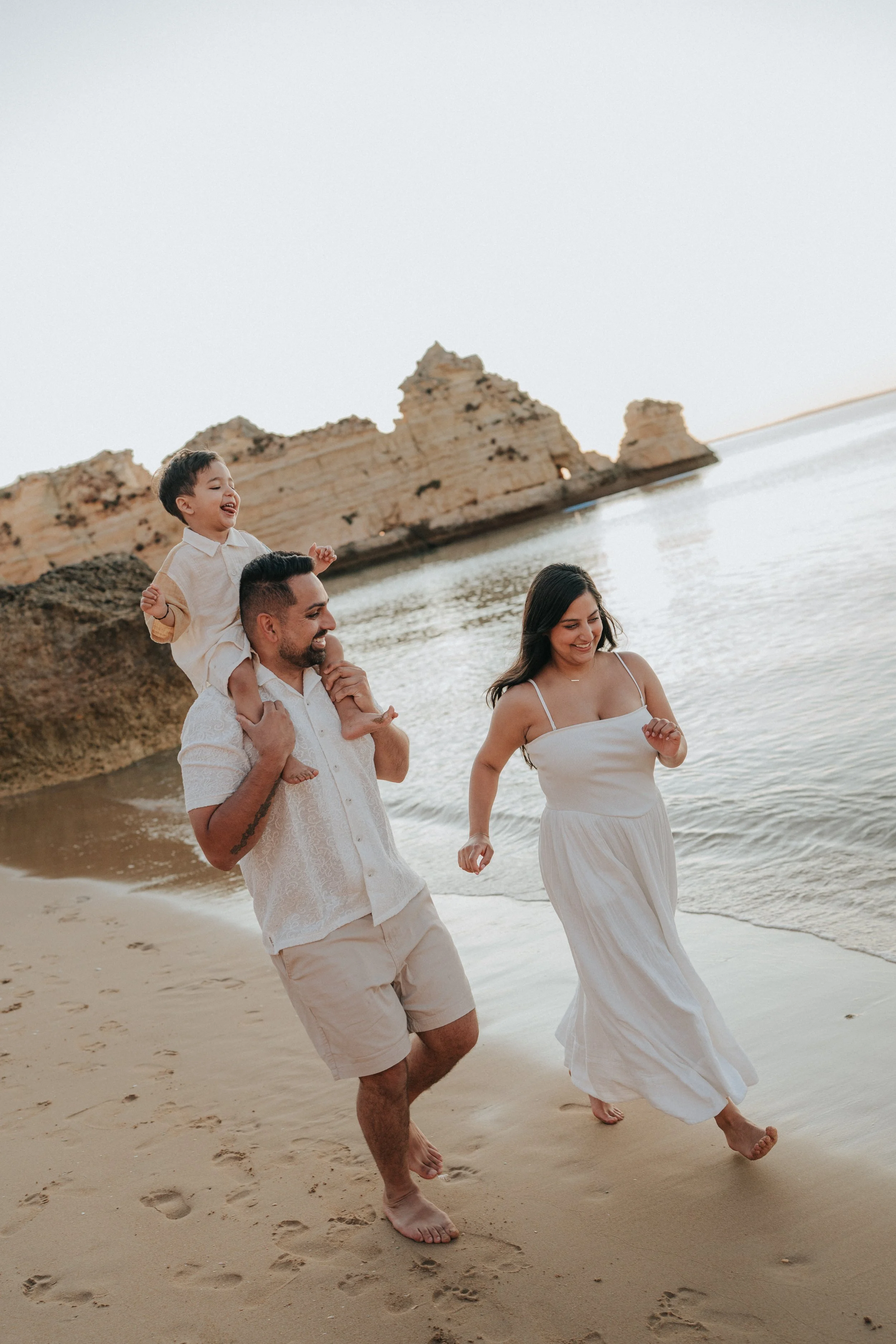 A family of three walking on the beach, with a rocky cliff in the background. The father is carrying a young boy on his shoulders, both smiling, and the mother is walking alongside them, also smiling.