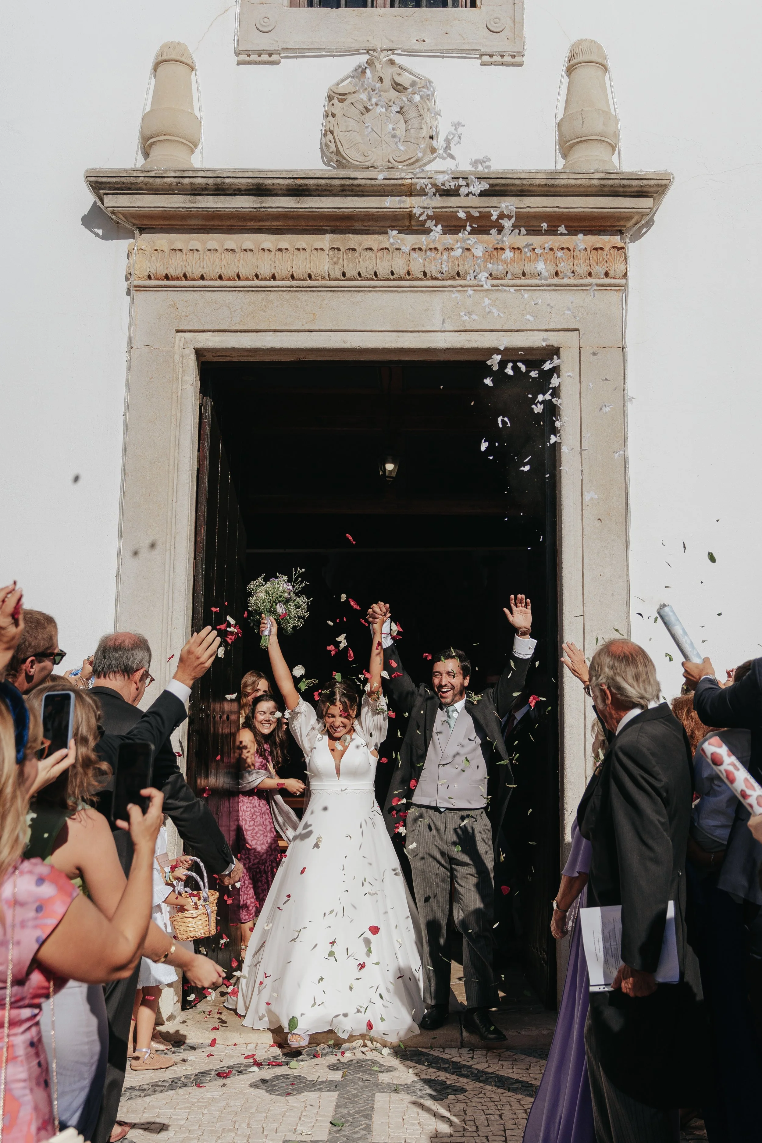 A bride and groom celebrate their wedding outside a building with guests surrounding them. The couple is smiling, with the bride holding a bouquet and the groom raising his hands. There are flower petals falling, and people are taking pictures and ch